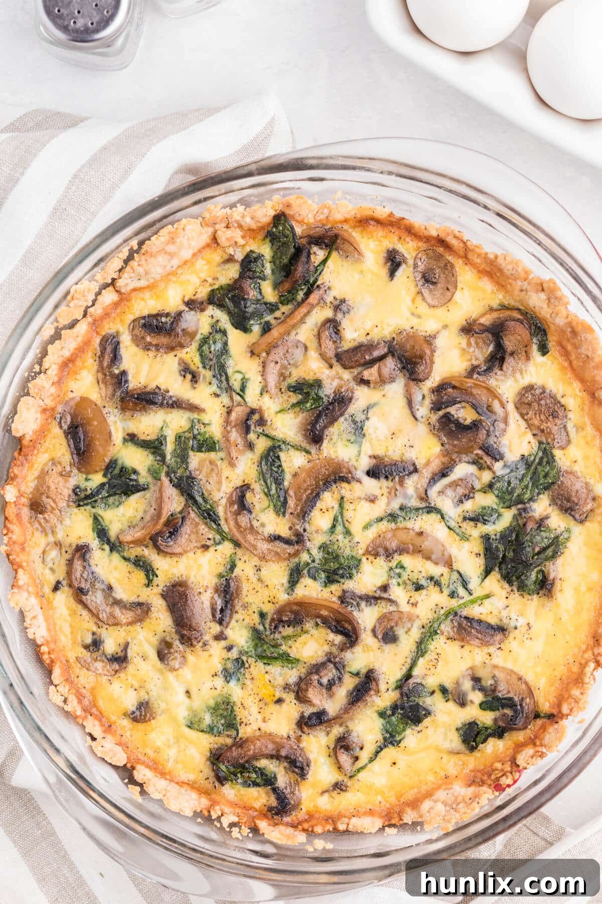 Earthy Mushroom and Fresh Spinach Quiche 2 A golden-brown Spinach Mushroom Quiche, freshly baked in a deep-dish pie pan, resting on a rustic wooden surface. The crust is perfectly browned, and the creamy filling, studded with green spinach and savory mushrooms, is visible through a single slice removed.