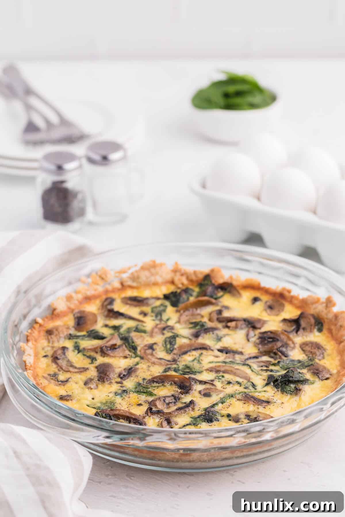 Earthy Mushroom and Fresh Spinach Quiche 7 A whole Spinach Mushroom Quiche in a ceramic pie pan, placed on a white and grey patterned towel on a wooden table. A single slice has been removed, revealing the creamy filling.
