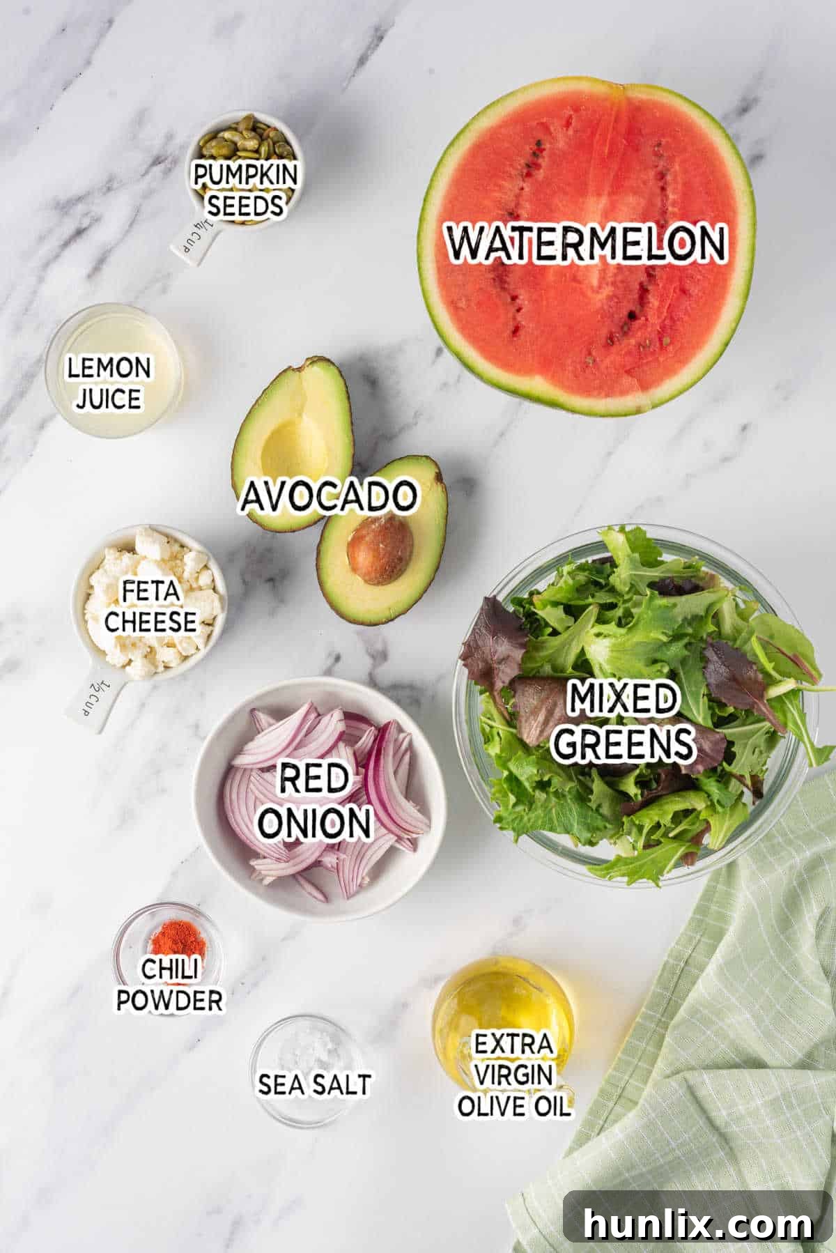 A vibrant display of fresh ingredients laid out, including a generous wedge of red watermelon, a block of creamy feta cheese, a ripe green avocado, a red onion, and a small bowl of pumpkin seeds, ready to be assembled for the Watermelon and Feta Salad.