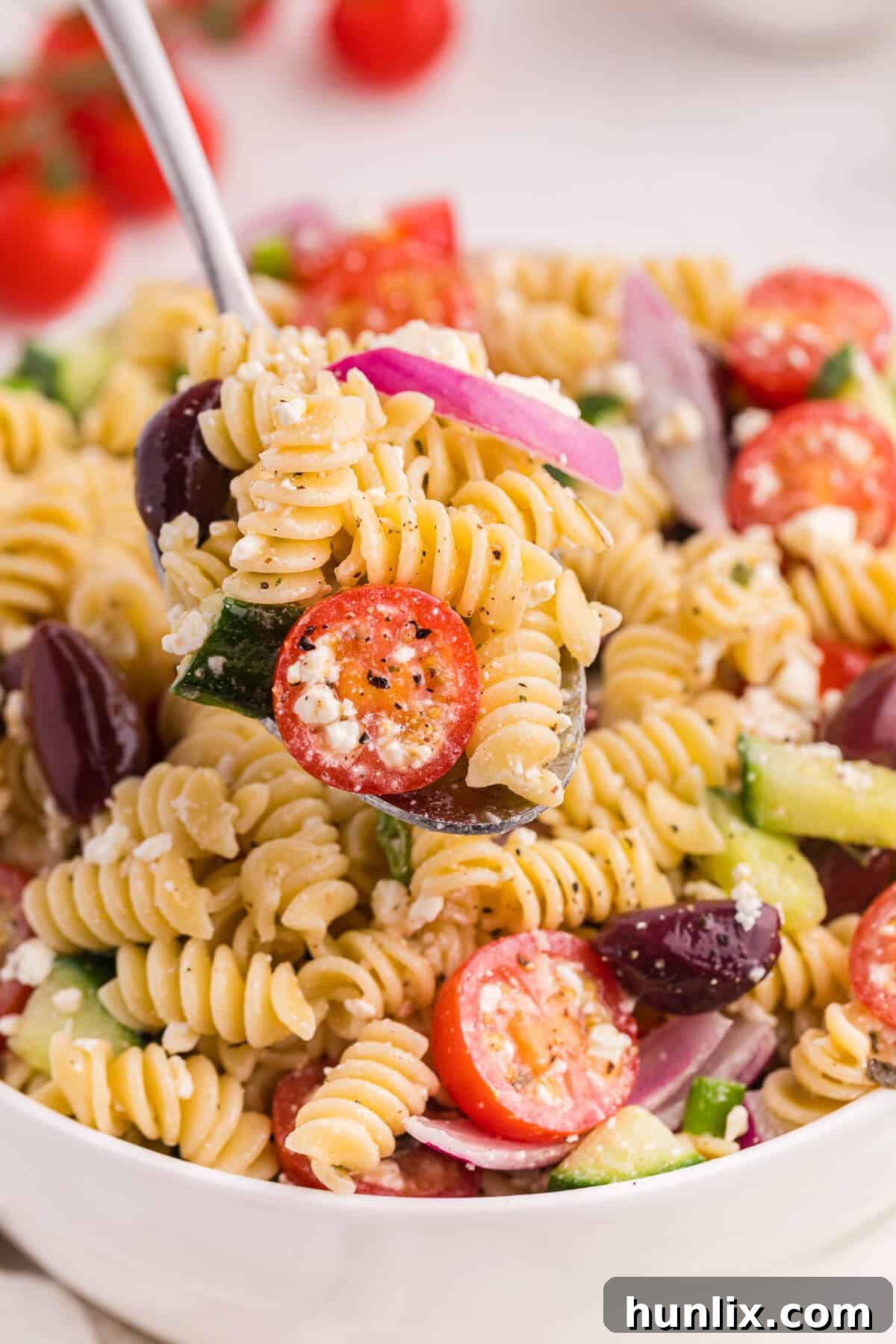 Zesty Greek Pasta Medley 6 A serving spoon lifting a portion of Greek pasta salad from a large bowl, highlighting the colorful mix of pasta, olives, tomatoes, cucumbers, and feta cheese, ready to be enjoyed.