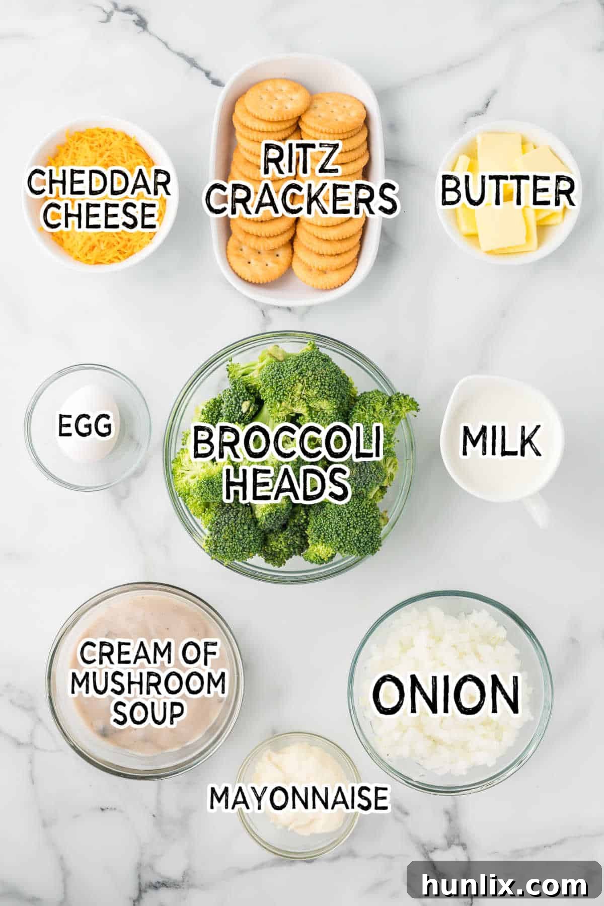 Fresh ingredients laid out on a white marble counter for making broccoli casserole, including broccoli heads, a yellow onion, cheddar cheese block, canned soup, milk, mayonnaise, eggs, Ritz crackers, and butter.