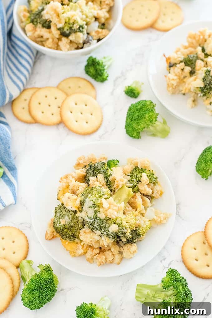 A serving of broccoli casserole on a white plate, surrounded by fresh broccoli florets and crackers on a wooden surface.