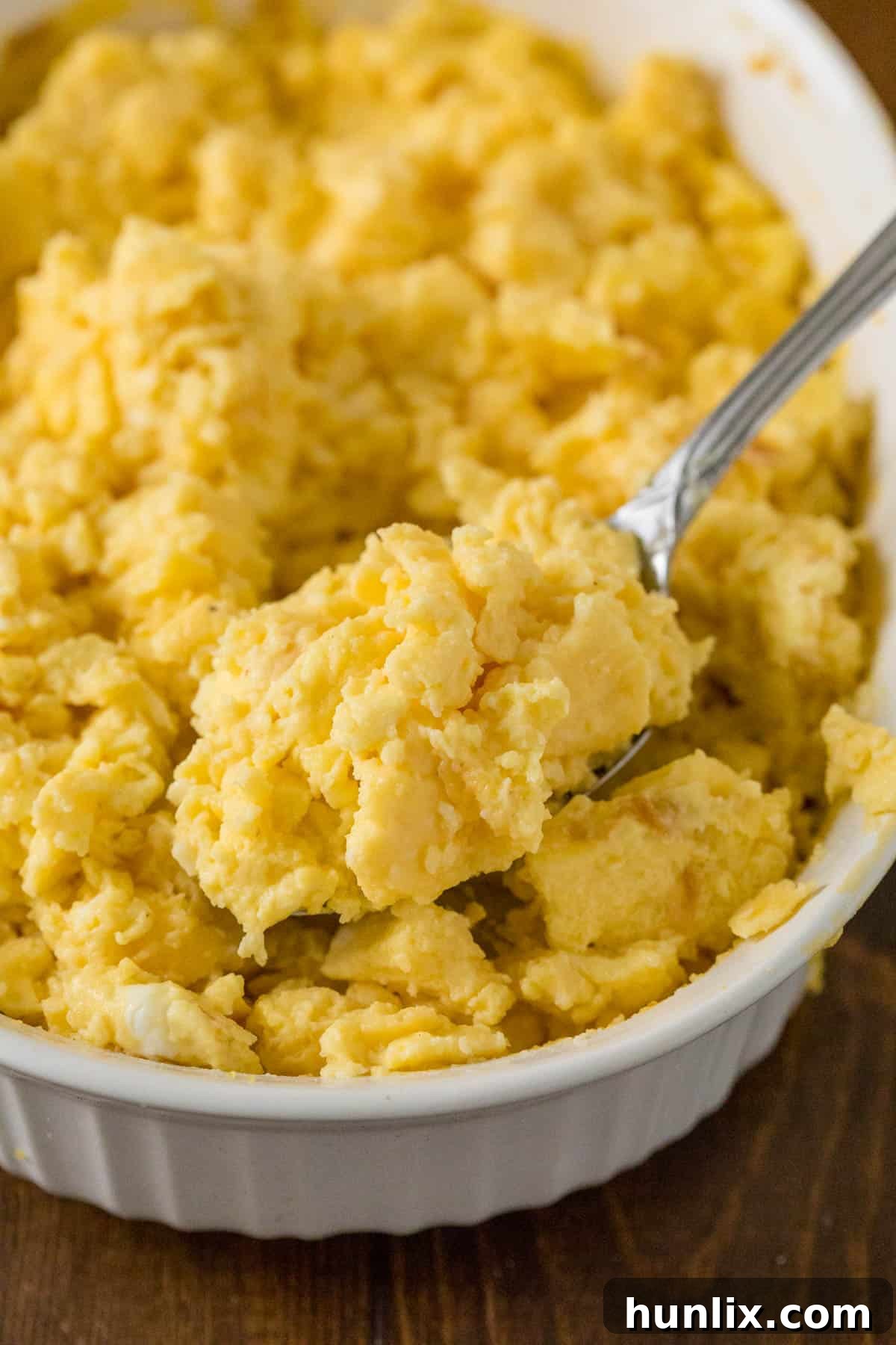 A close-up of oven scrambled eggs on a serving spoon, lifted from a casserole dish, highlighting their fluffy texture.