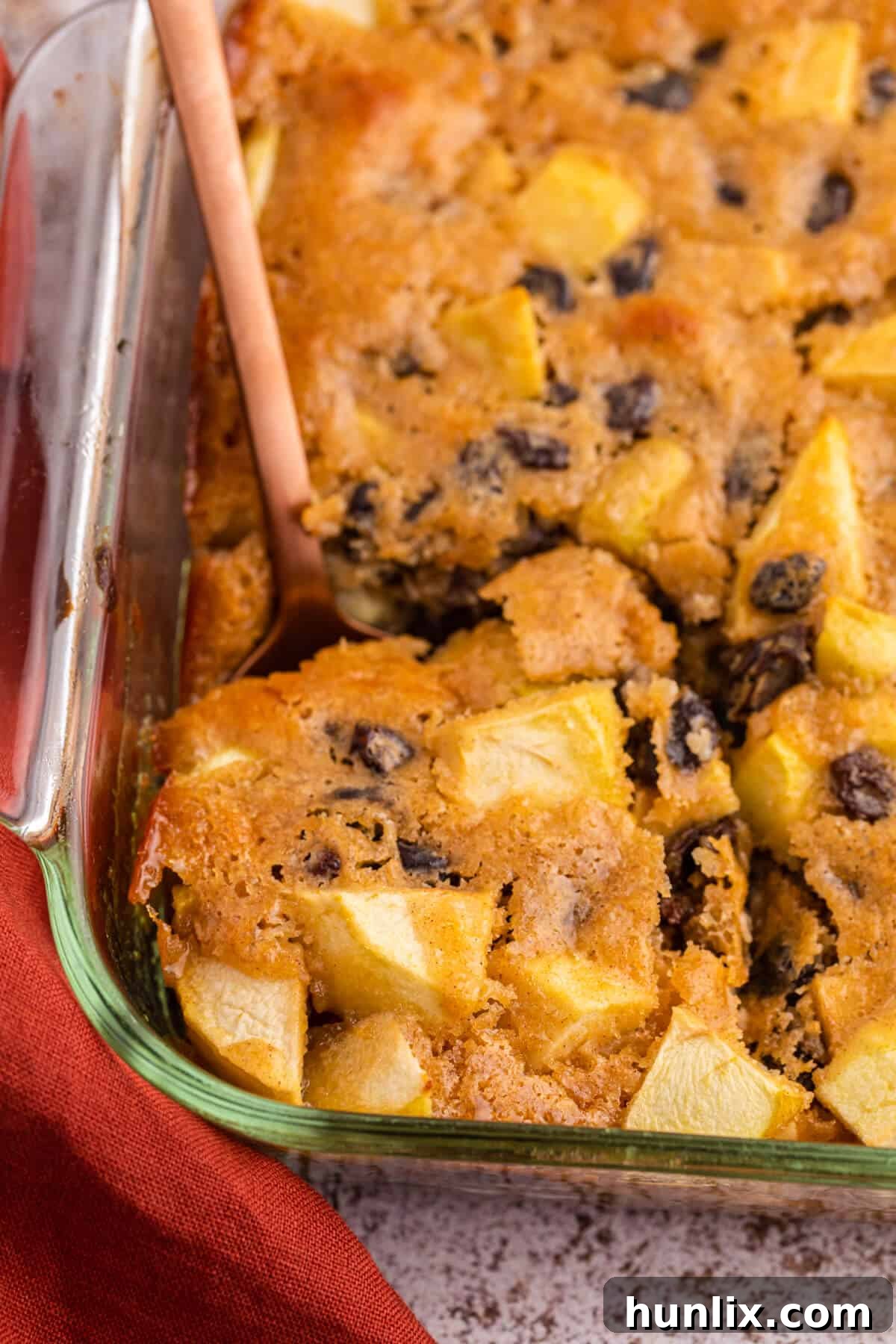 A serving spoon lifting a piece of warm maple apple pudding from a baking dish, highlighting its rich, custardy texture.