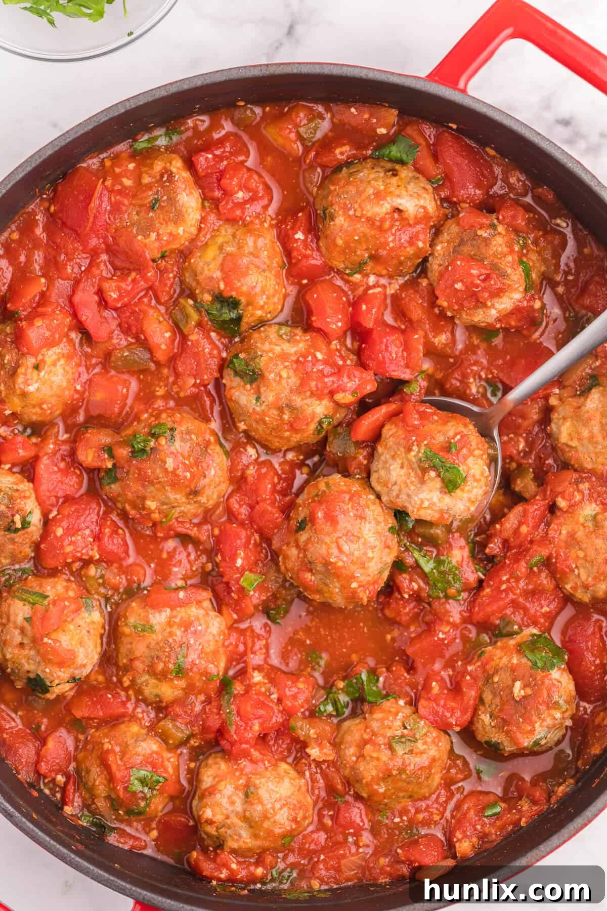 A close-up of Mexican meatballs in a red skillet with a serving spoon, garnished with fresh cilantro, ready to be dished out.