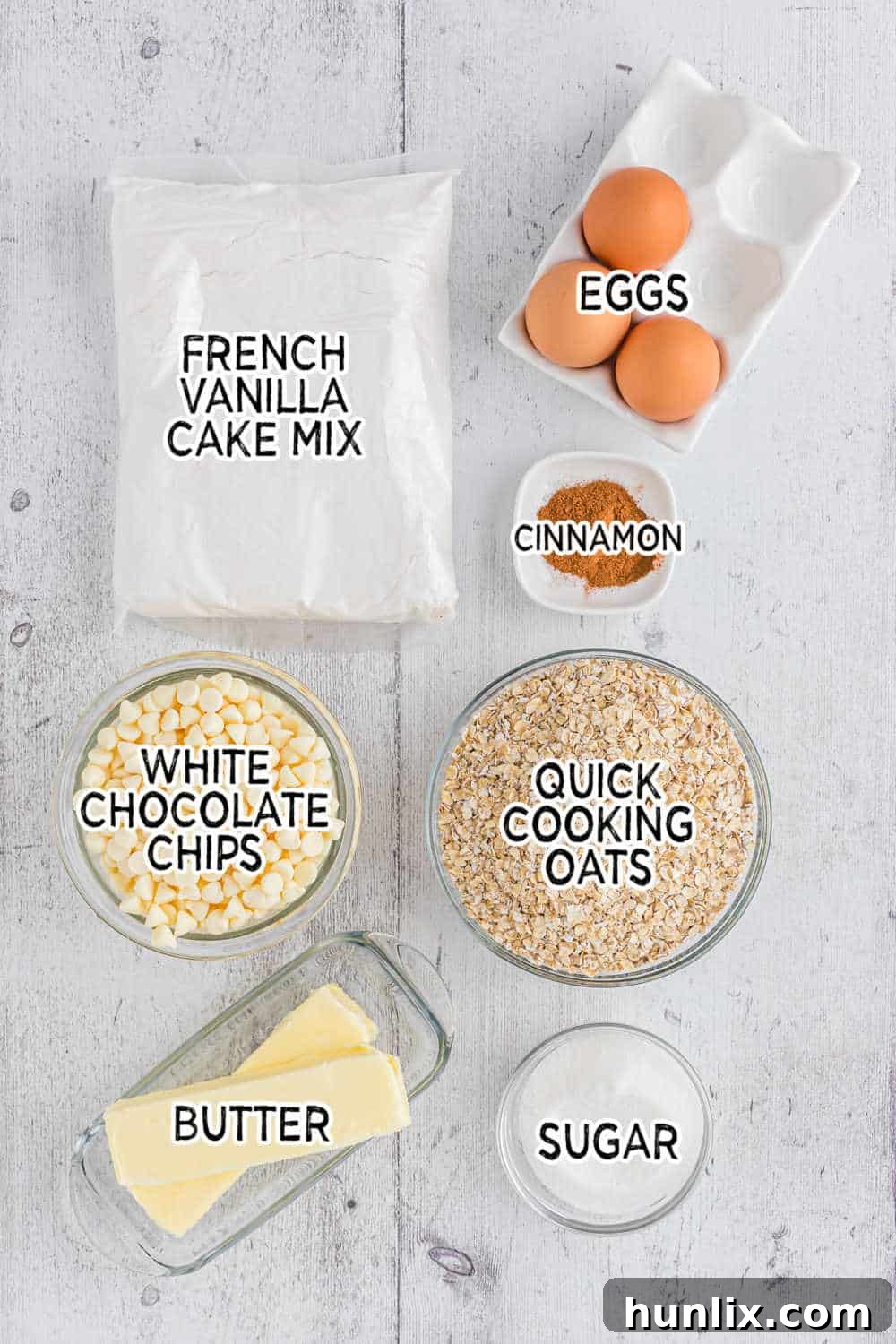 Various ingredients for white chocolate oatmeal cookies laid out on a surface, including butter, sugar, cinnamon, eggs, cake mix, quick oats, and a bag of white chocolate chips.