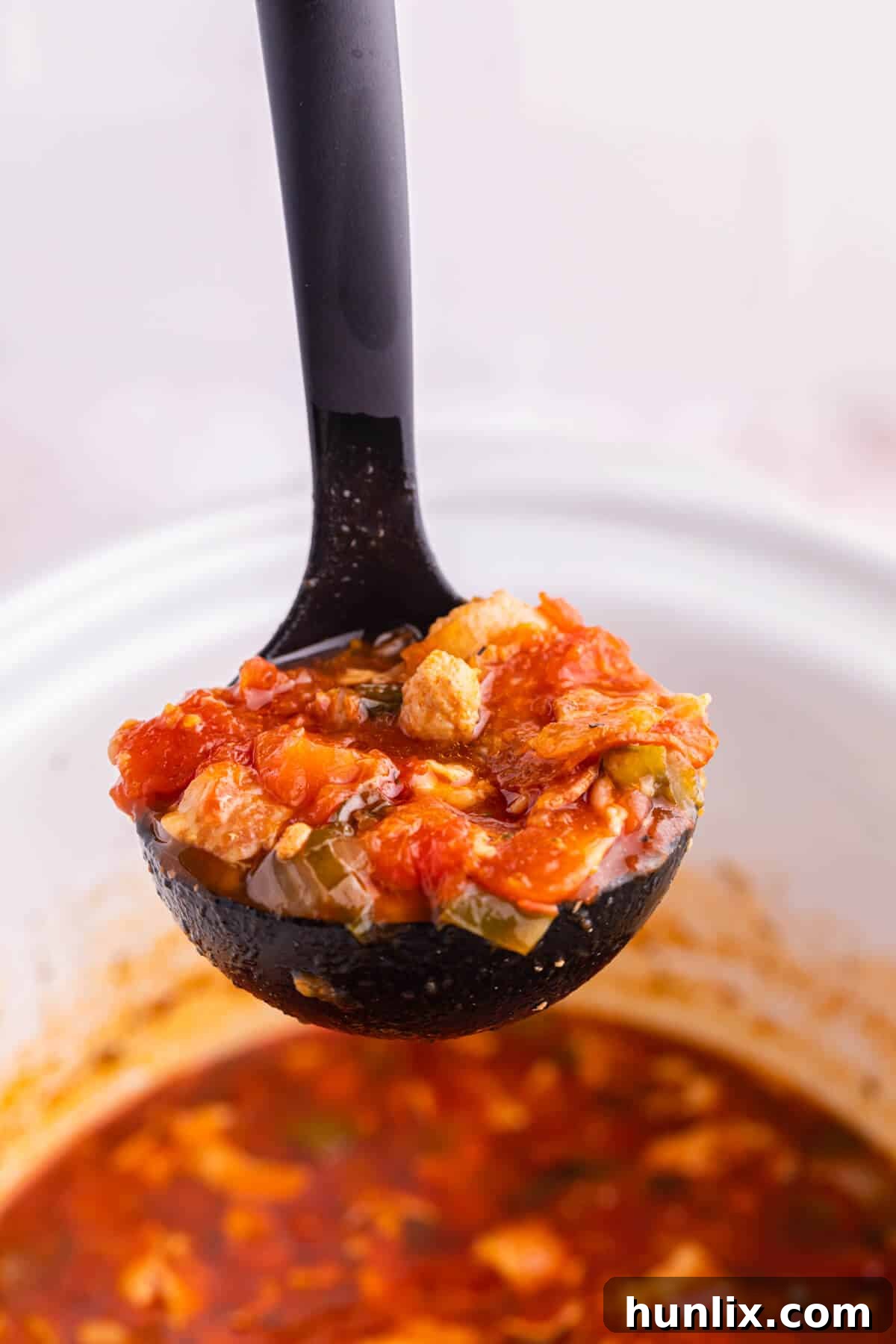 A close-up of a ladle full of rich pizza soup, held over a white slow cooker.