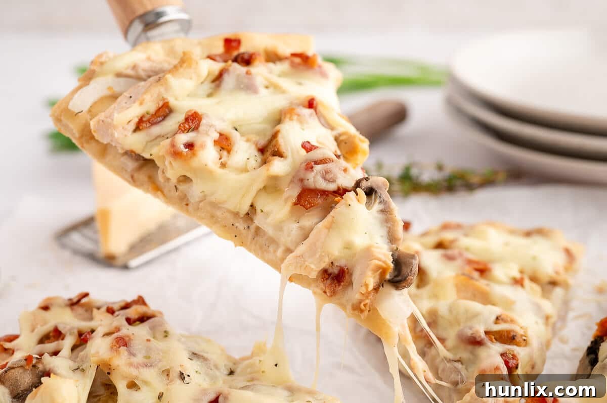A perfect slice of chicken bacon alfredo pizza being lifted from the whole pizza with a spatula, showcasing its creamy sauce, melted cheese, and generous toppings.