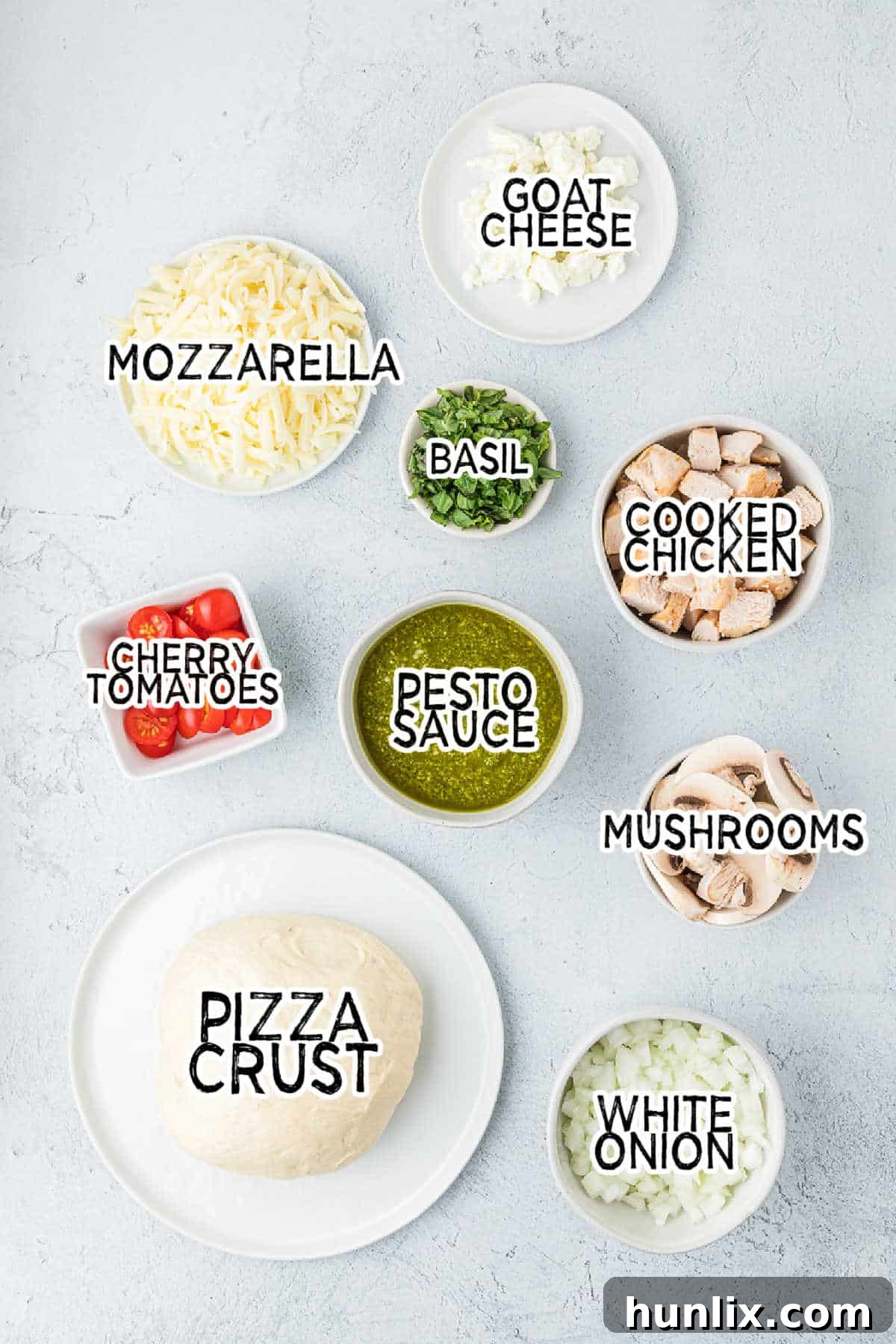 Various fresh ingredients for chicken pesto pizza, including pizza dough, pesto sauce, chicken, vegetables, and cheeses, arranged on a kitchen countertop.