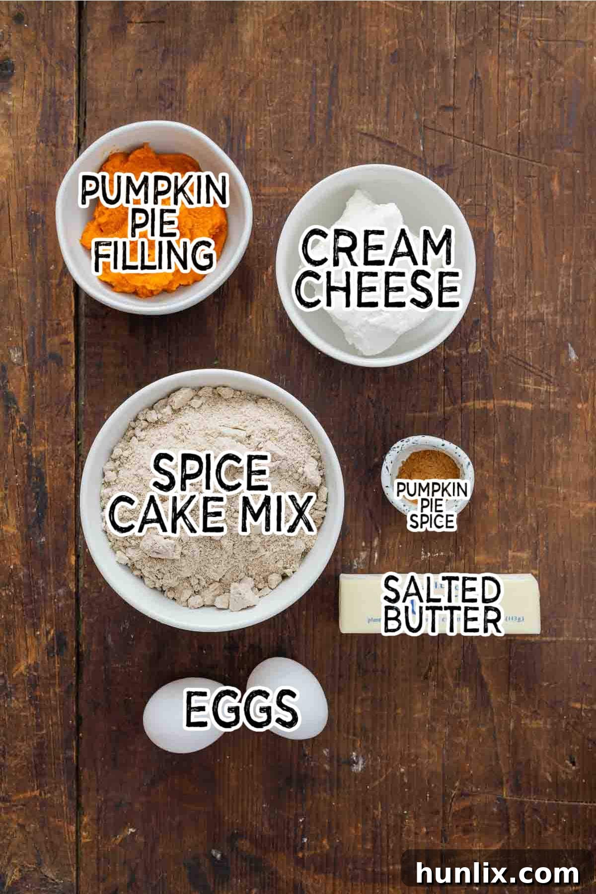 Autumn's Embrace Cakelets 3 Various ingredients for pumpkin spice cake cups, including a box of spice cake mix, eggs, butter, cream cheese, pumpkin pie filling, and a small bowl of pumpkin pie spice, neatly arranged in white bowls on a rustic wooden surface.
