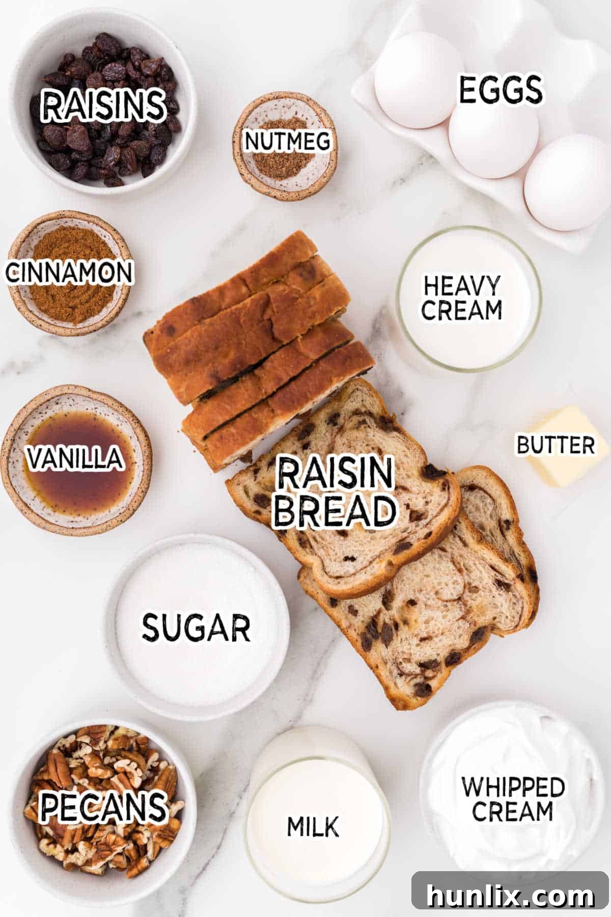 A collection of ingredients for cinnamon raisin bread pudding neatly arranged on a countertop, including a loaf of bread, eggs, milk, cream, spices, raisins, and pecans.