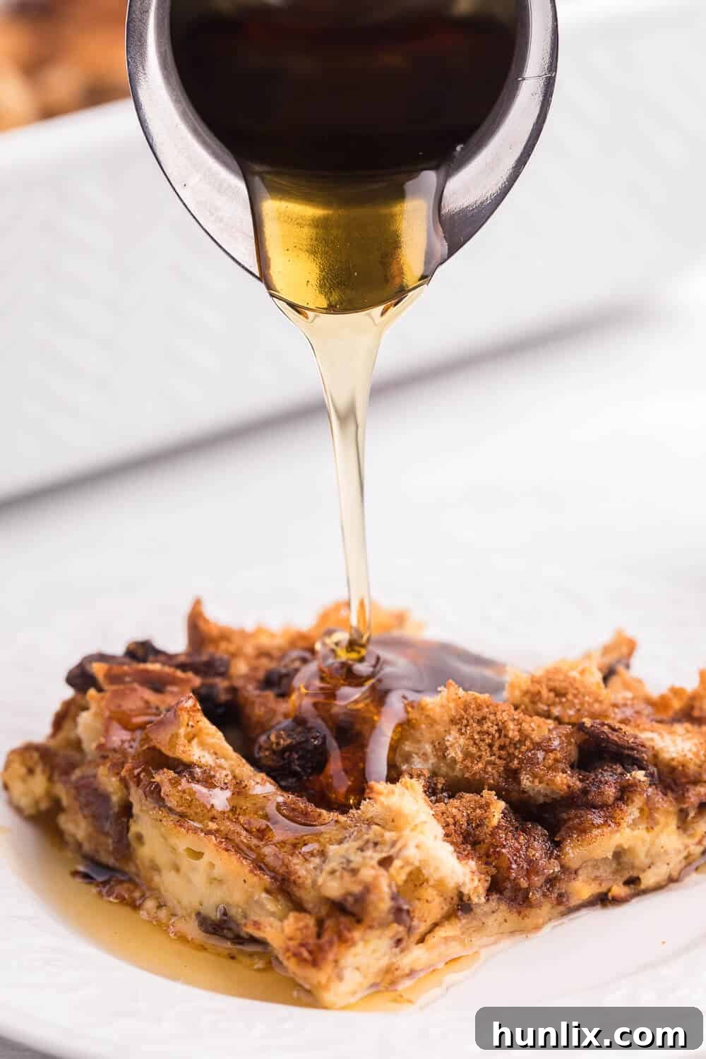 Maple syrup being poured over a slice of Cinnamon Raisin Breakfast Casserole on a plate, adding a luscious finish.