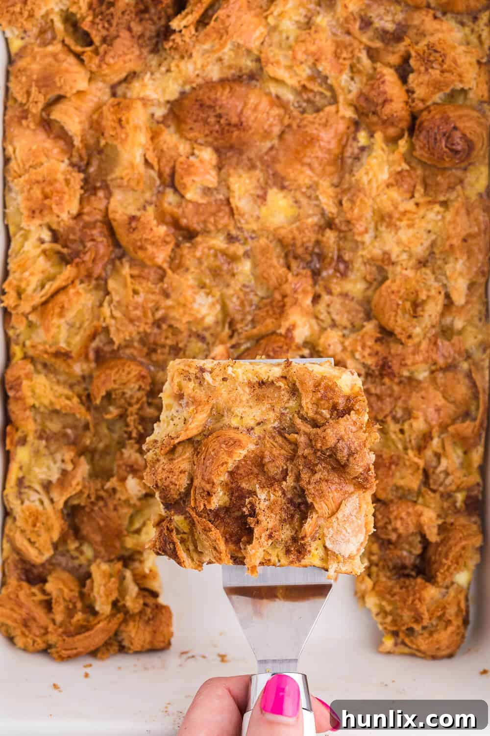 A spatula lifting a slice of eggnog breakfast casserole from a white baking dish, showing its creamy layers.