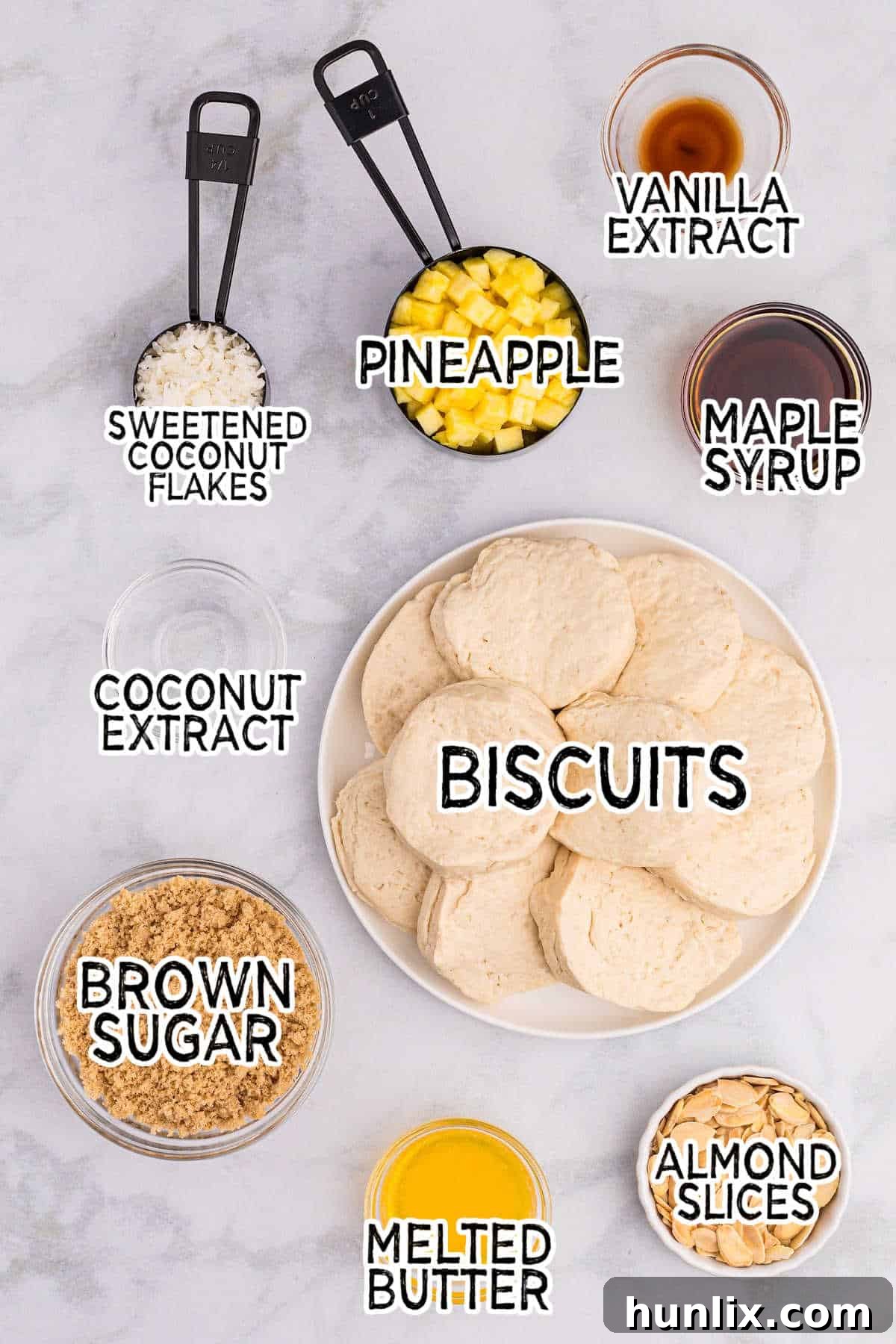 Pineapple Paradise Sticky Buns with Coconut Caramel Sauce 3 Various ingredients laid out on a white surface, including a can of biscuits, brown sugar, maple syrup, melted butter, vanilla extract, coconut extract, chopped pineapple, sliced almonds, and sweetened coconut flakes, ready for making pineapple sticky buns.