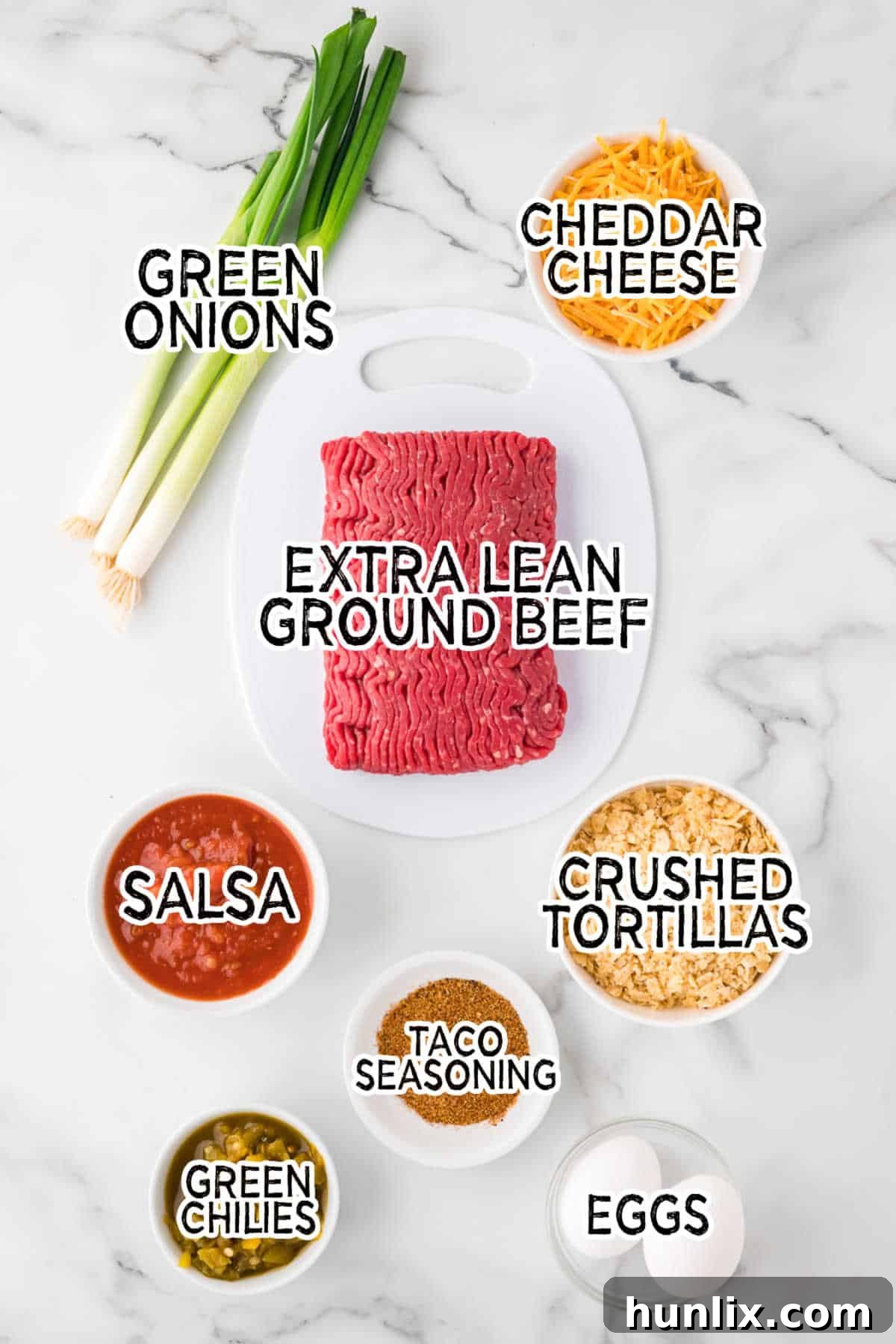All the fresh ingredients for Mexican Meatloaf laid out on a white marble background, including ground beef, crushed tortilla chips, cheese, salsa, eggs, taco seasoning, green chilies, and green onions.