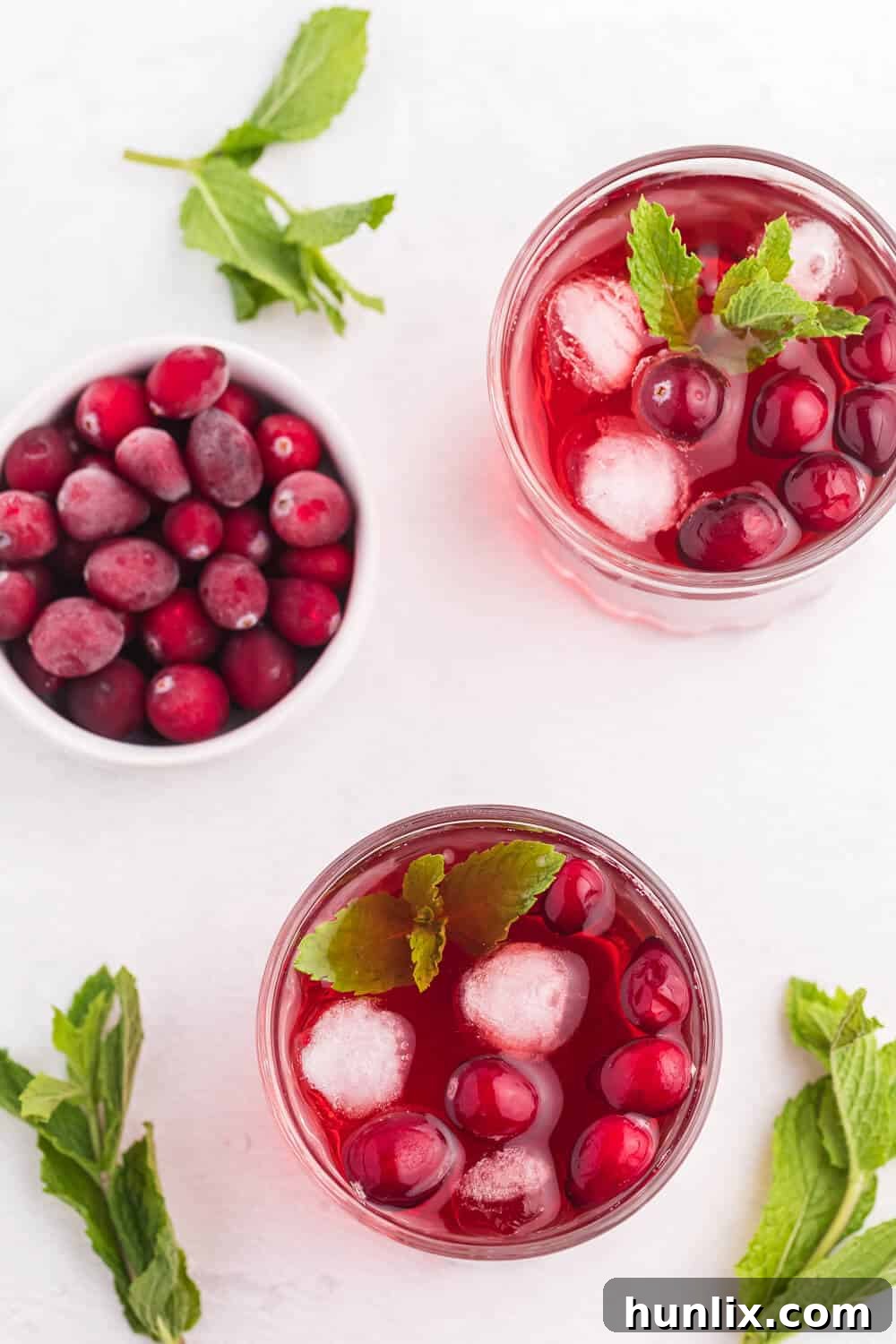 Glasses of the Cranberry Mint Mocktail on a white surface with mint leaves scattered around.