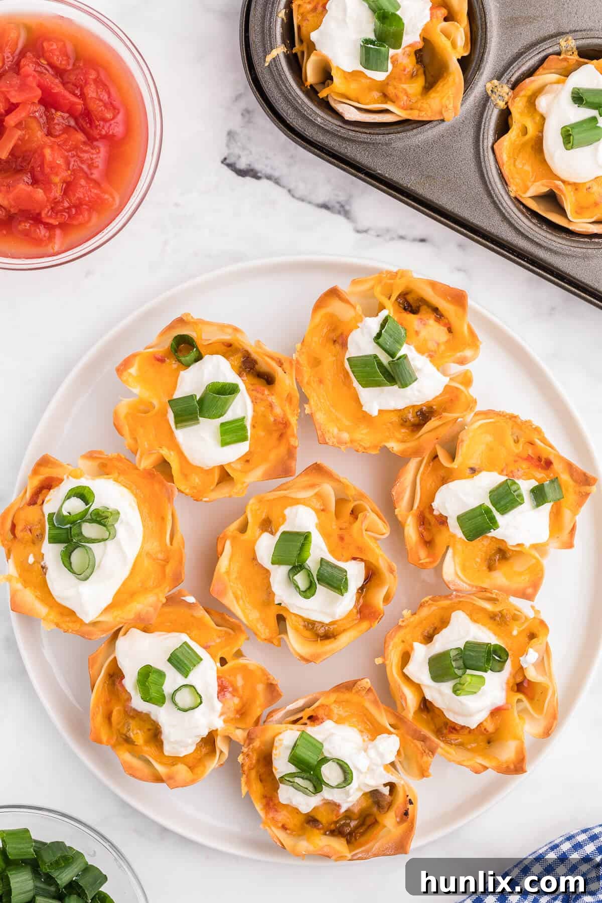 Freshly baked Taco Wonton Cups cooling slightly in a muffin tin, with some already topped with sour cream and green onions, ready to be served as an easy appetizer.