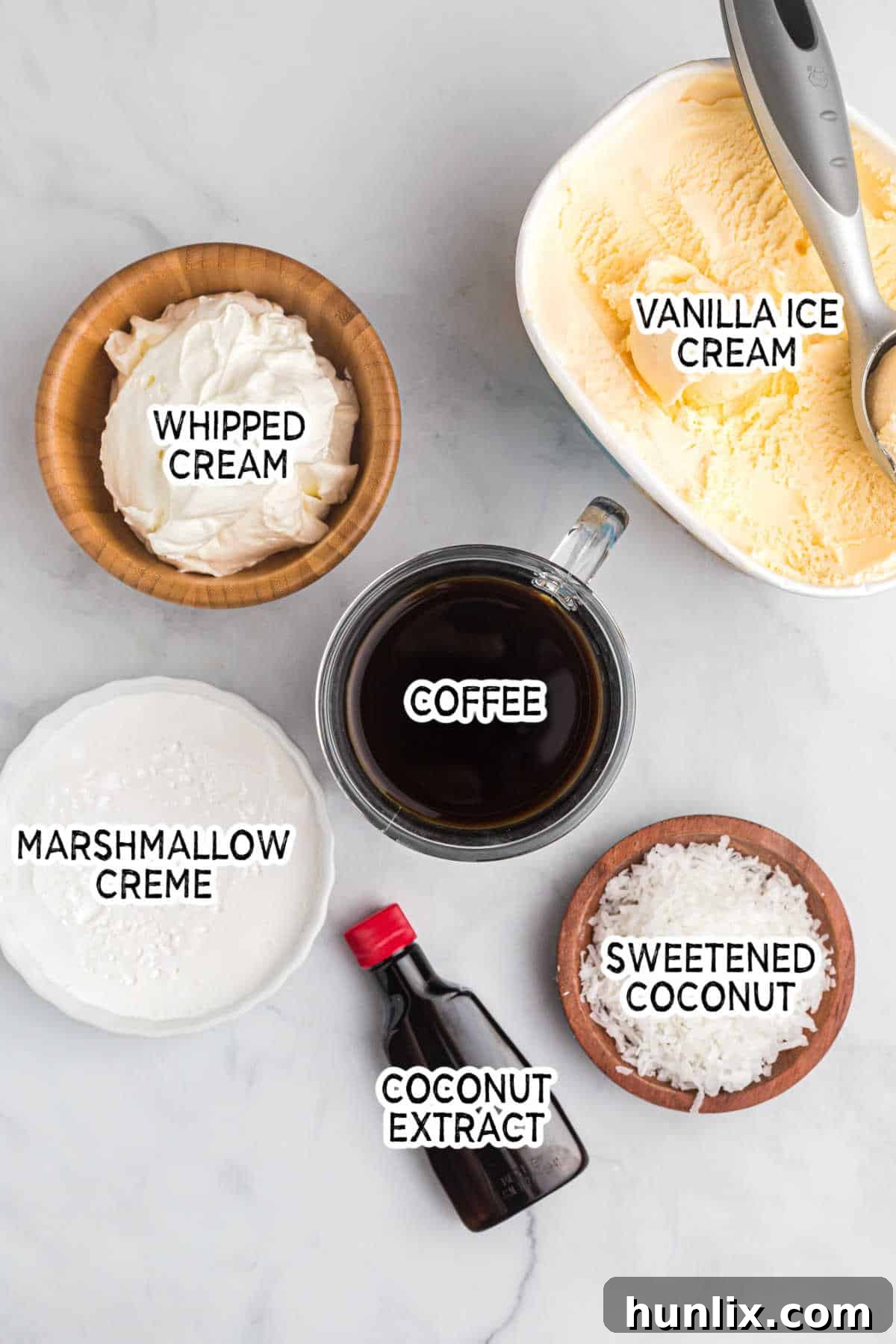 A collection of ingredients for coconut coffee milkshake, including a bag of shredded coconut, a container of marshmallow creme, a bottle of coconut extract, and a glass of brewed coffee, all arranged on a grey surface.