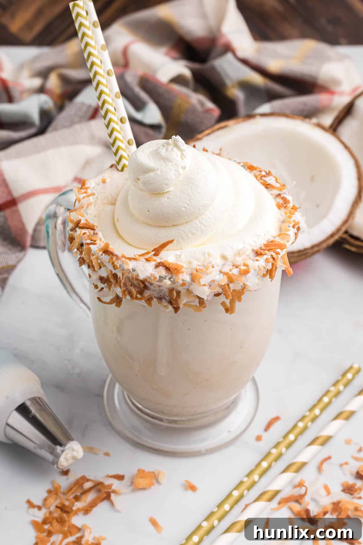 A tall glass of Coconut Coffee Milkshake with a straw, garnished with whipped cream and toasted coconut, set against a blurred background.