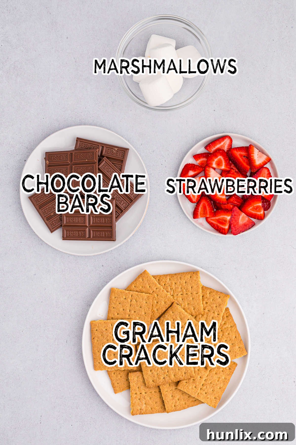 Various ingredients for strawberry s'mores laid out on a grey background, including graham crackers, strawberries, marshmallows, and chocolate bars.