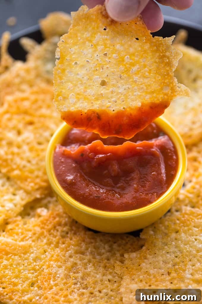 A hand reaching for a crispy cheese chip to dip it into a bowl of vibrant red pizza sauce, highlighting the snack's irresistible appeal.