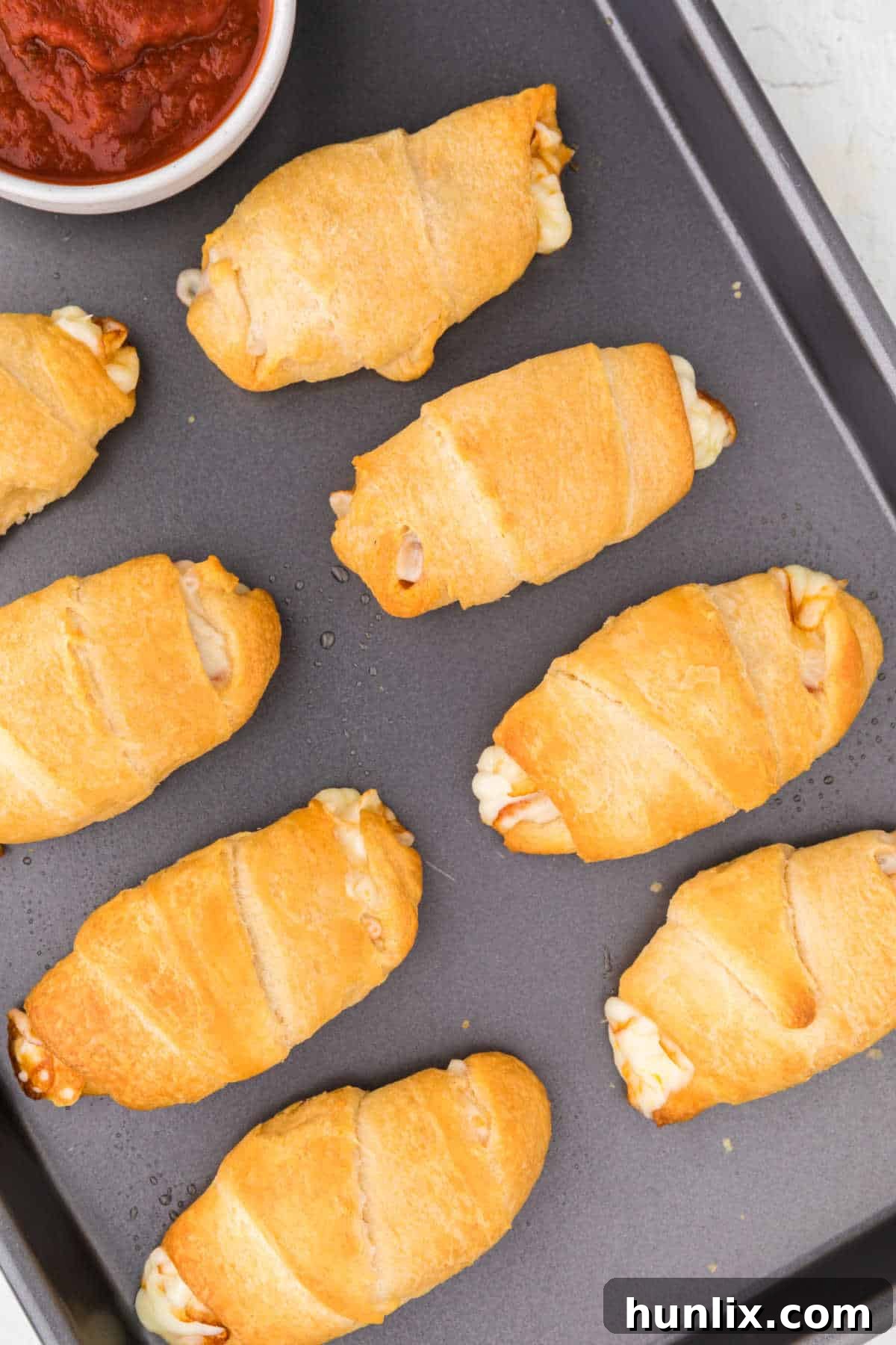 Golden brown pizza rolls on a sheet pan, ready to serve, with a small bowl of rich red pizza dipping sauce on the side.