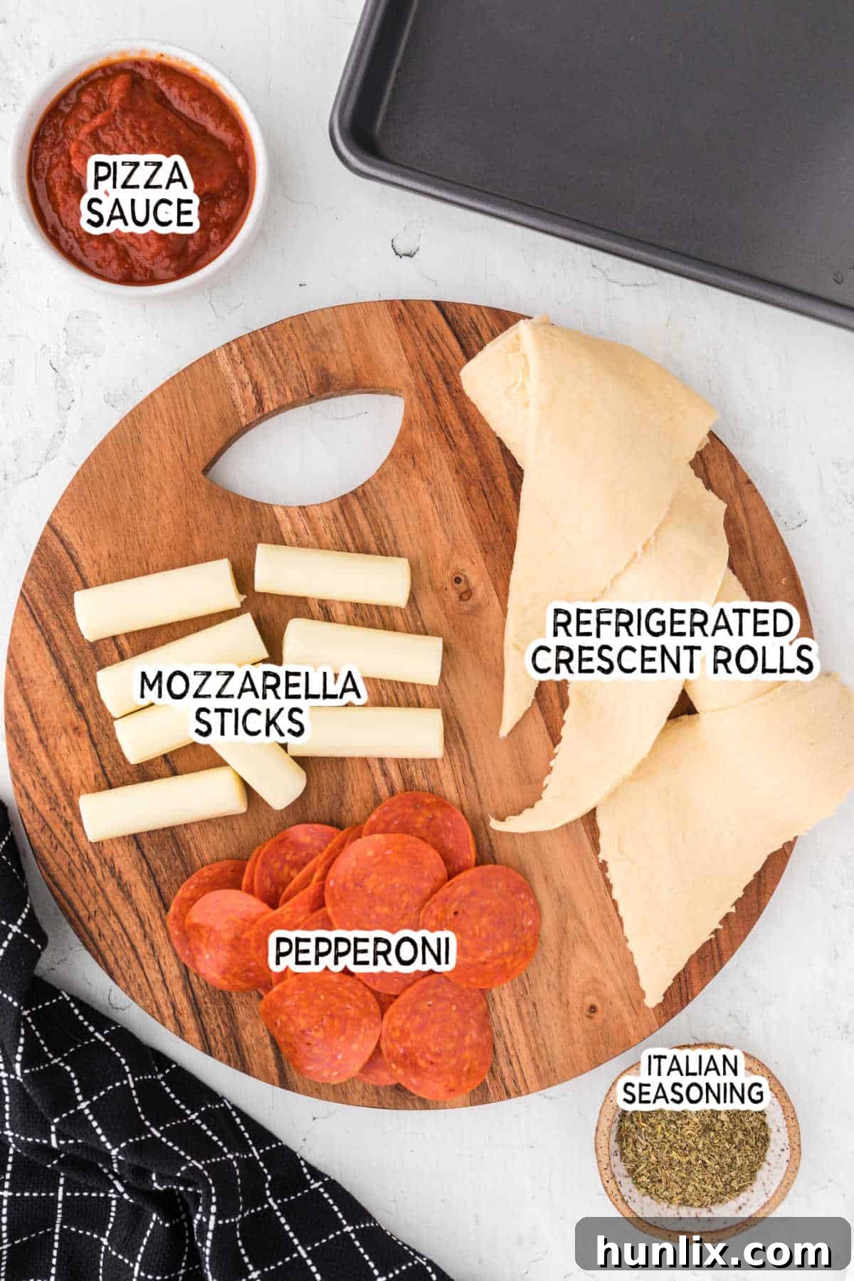 Ingredients for pizza rolls laid out on a round wooden cutting board: crescent roll dough, pepperoni slices, mozzarella sticks, and a bowl of pizza sauce.