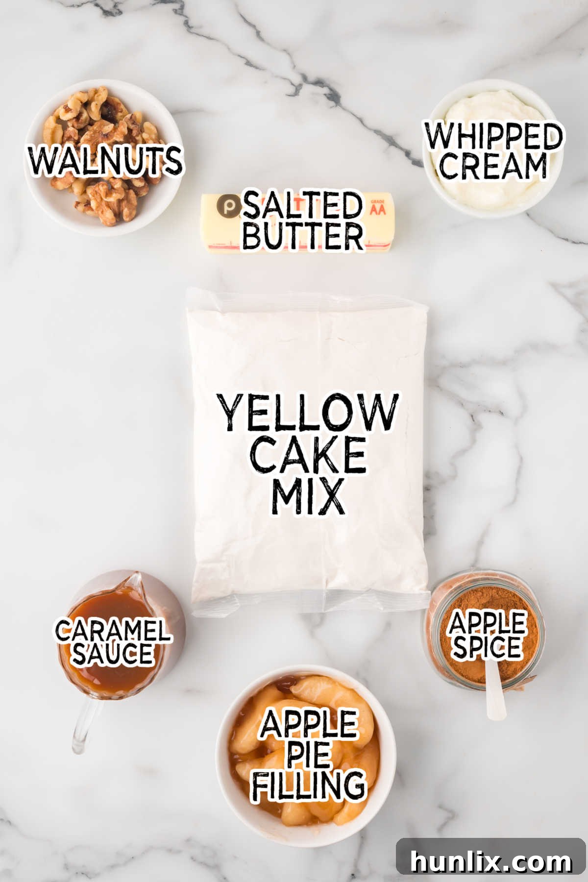 Various ingredients for an apple dump cake, including a can of apple pie filling, a jar of caramel sauce, a box of yellow cake mix, walnuts, butter sticks, and a container of apple spice, neatly arranged on a white marble counter.