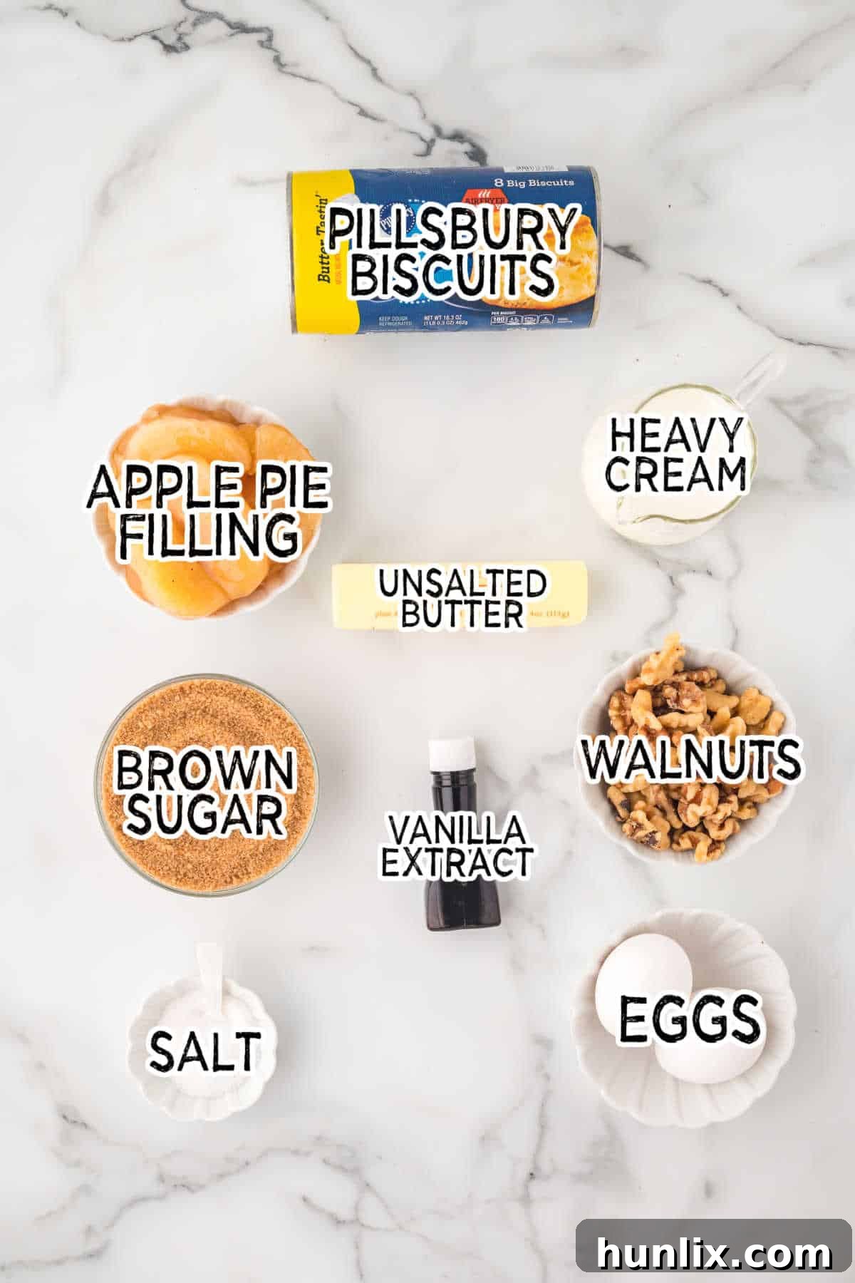 Individual ingredients for Apple Butterscotch Buns laid out on a white countertop: Pillsbury rolls, eggs, apple pie filling, butter, brown sugar, salt, heavy cream, vanilla extract, and walnuts.