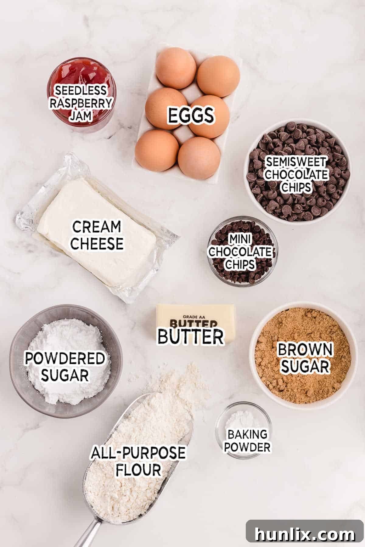 A collection of fresh ingredients laid out on a white surface, including butter, chocolate chips, eggs, brown sugar, flour, baking powder, cream cheese, and raspberry jam.