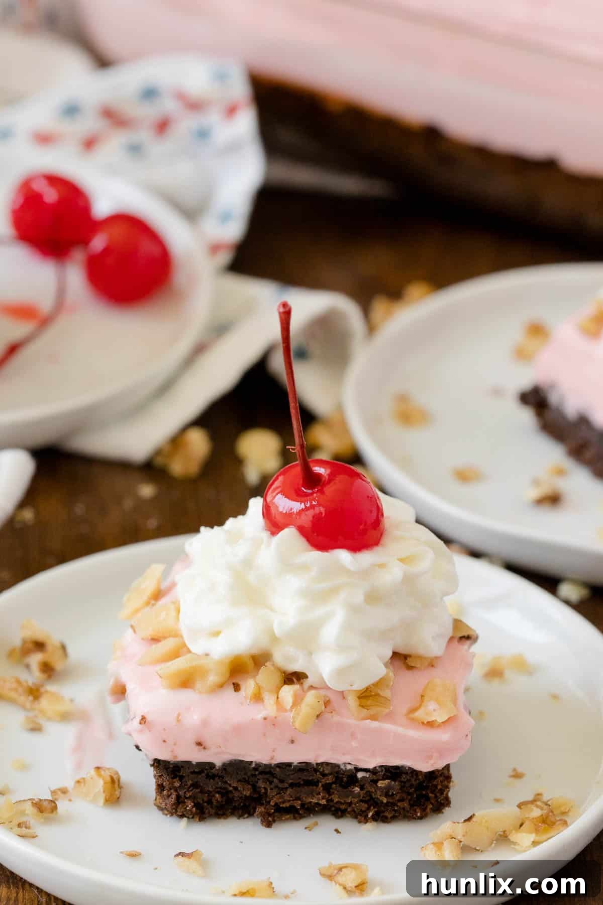 A perfectly cut square of Banana Split Brownie, topped with pink frosting and a cherry, resting on a white plate, ready to be enjoyed.