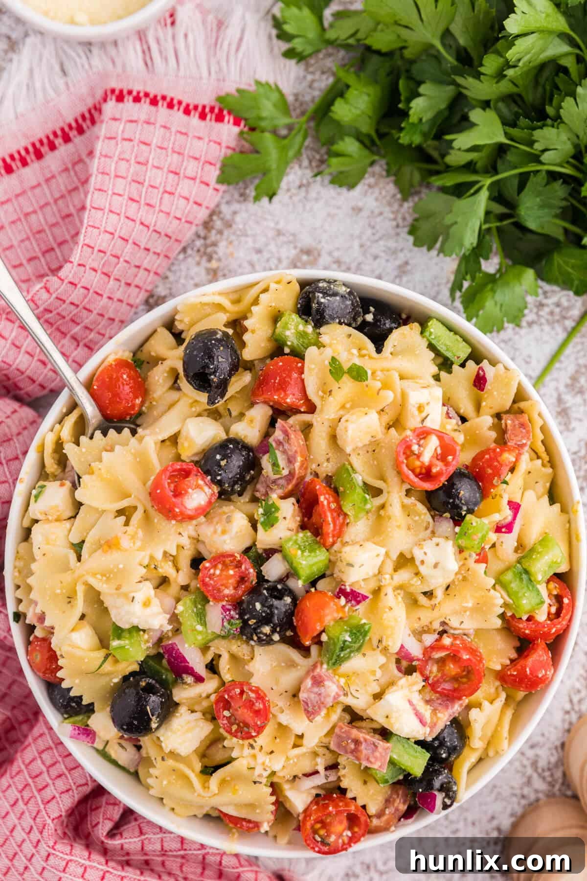 A large bowl filled with colorful Italian pasta salad, featuring bowtie pasta, red and green vegetables, olives, and salami. A serving spoon rests in the bowl.