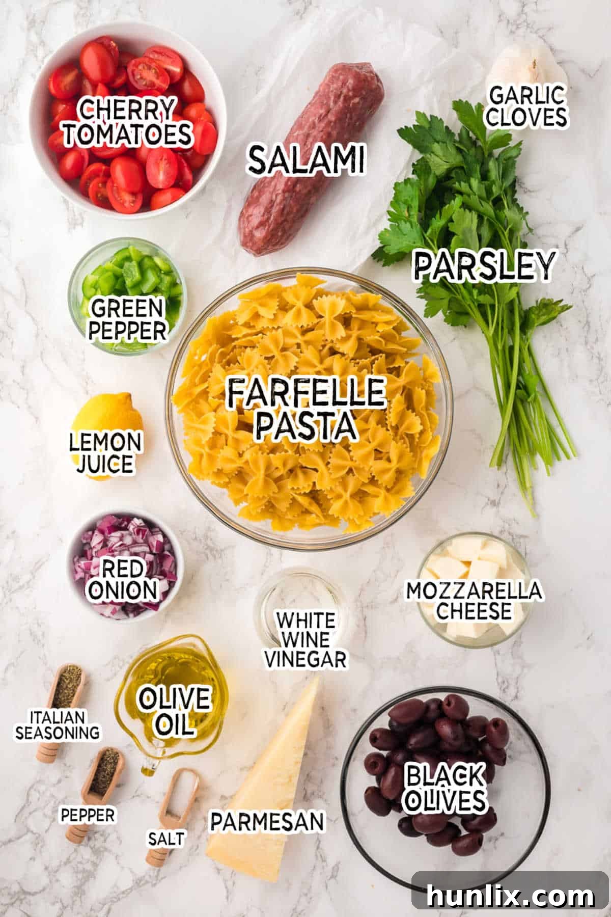 Ingredients for Italian Pasta Salad arranged on a white marble countertop, including uncooked pasta, fresh vegetables, salami, olives, and dressing components.