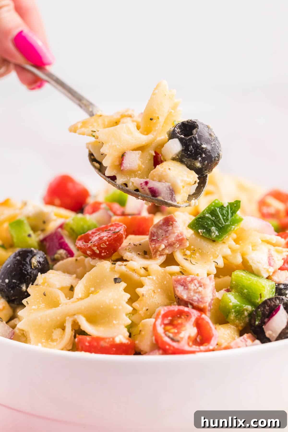 A close-up of a spoon holding a portion of Italian pasta salad, showing the vibrant mixture of pasta, vegetables, cheese, and salami.