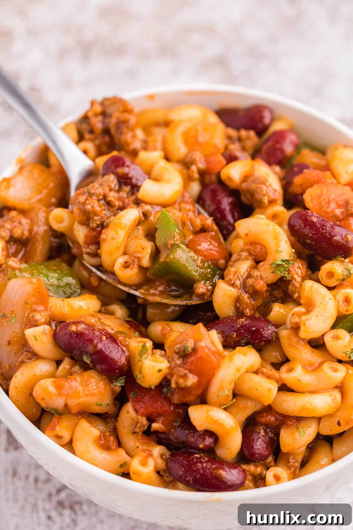 A white bowl filled with Chili Macaroni and a spoon, ready to be eaten.
