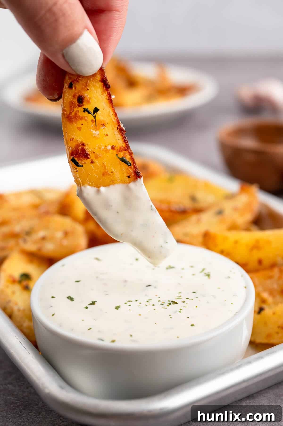 A hand reaching to dip a Parmesan thyme roasted potato wedge into a small bowl of creamy ranch dip.