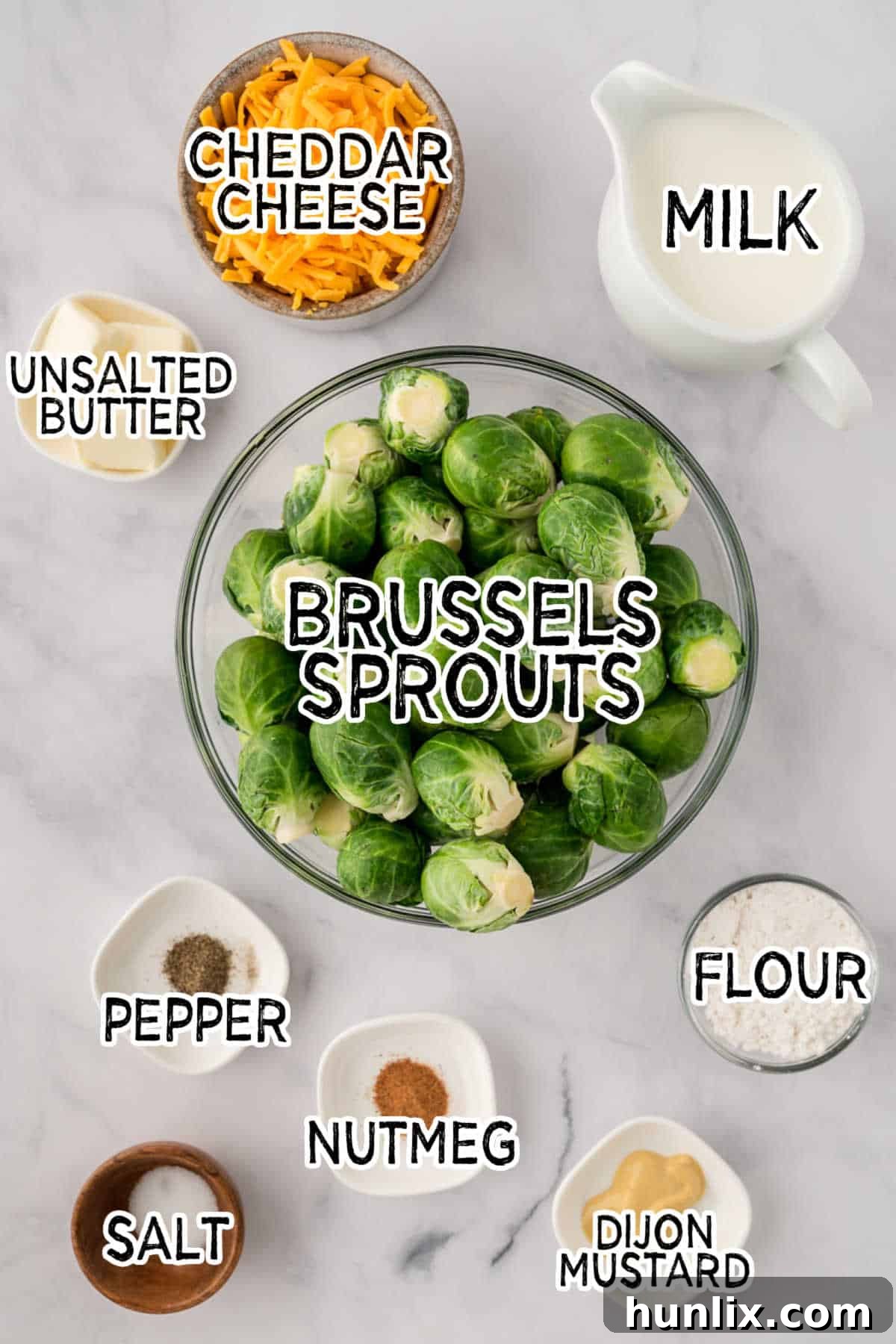 A carefully arranged spread of ingredients on a white marble counter, including fresh Brussels sprouts, butter, flour, milk, Dijon mustard, and a block of cheddar cheese.