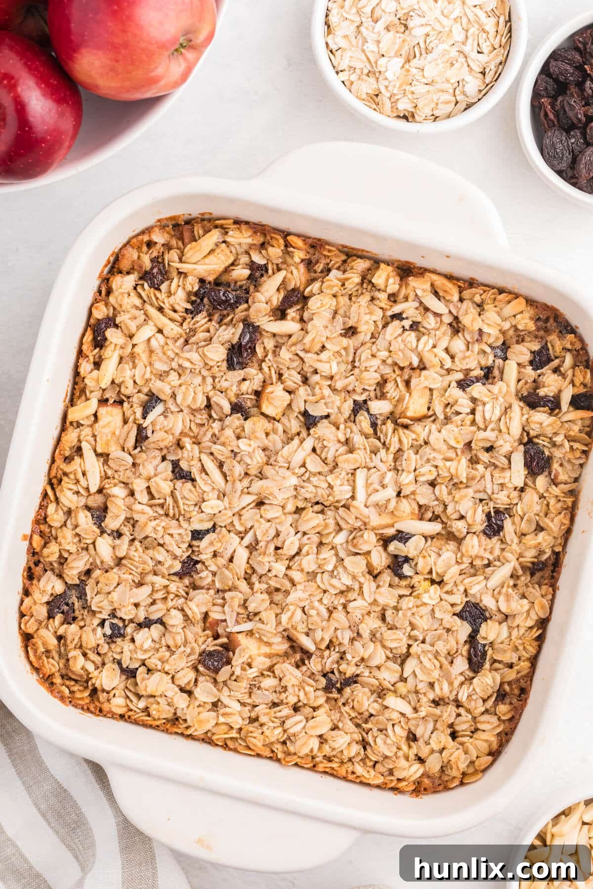 Baked oatmeal in a casserole pan, adorned with fresh apple slices, plump raisins, and crunchy slivered almonds. The golden-brown surface hints at a perfectly baked, comforting breakfast.