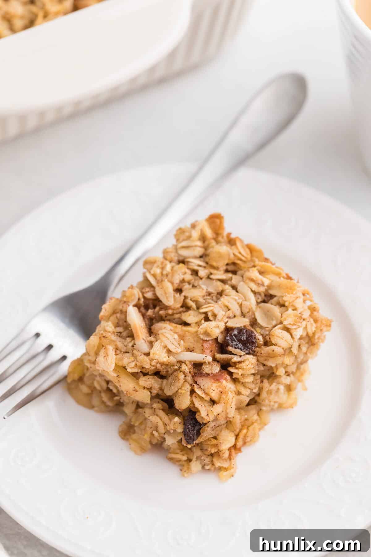 A perfectly portioned slice of baked oatmeal on a white plate, served with a fork, showing its hearty texture and delicious layers of fruit and nuts.