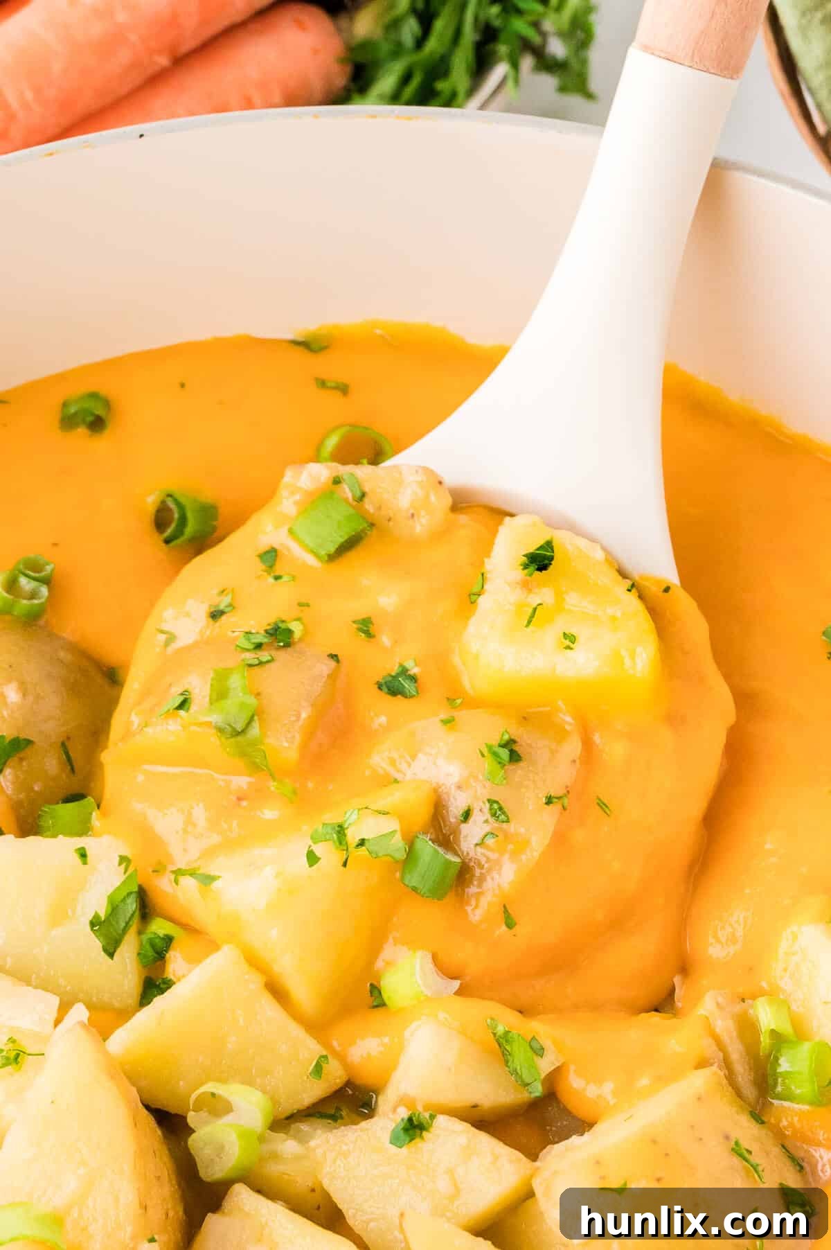 A white ceramic ladle brimming with smooth, orange carrot and potato soup, held above a rustic pot.