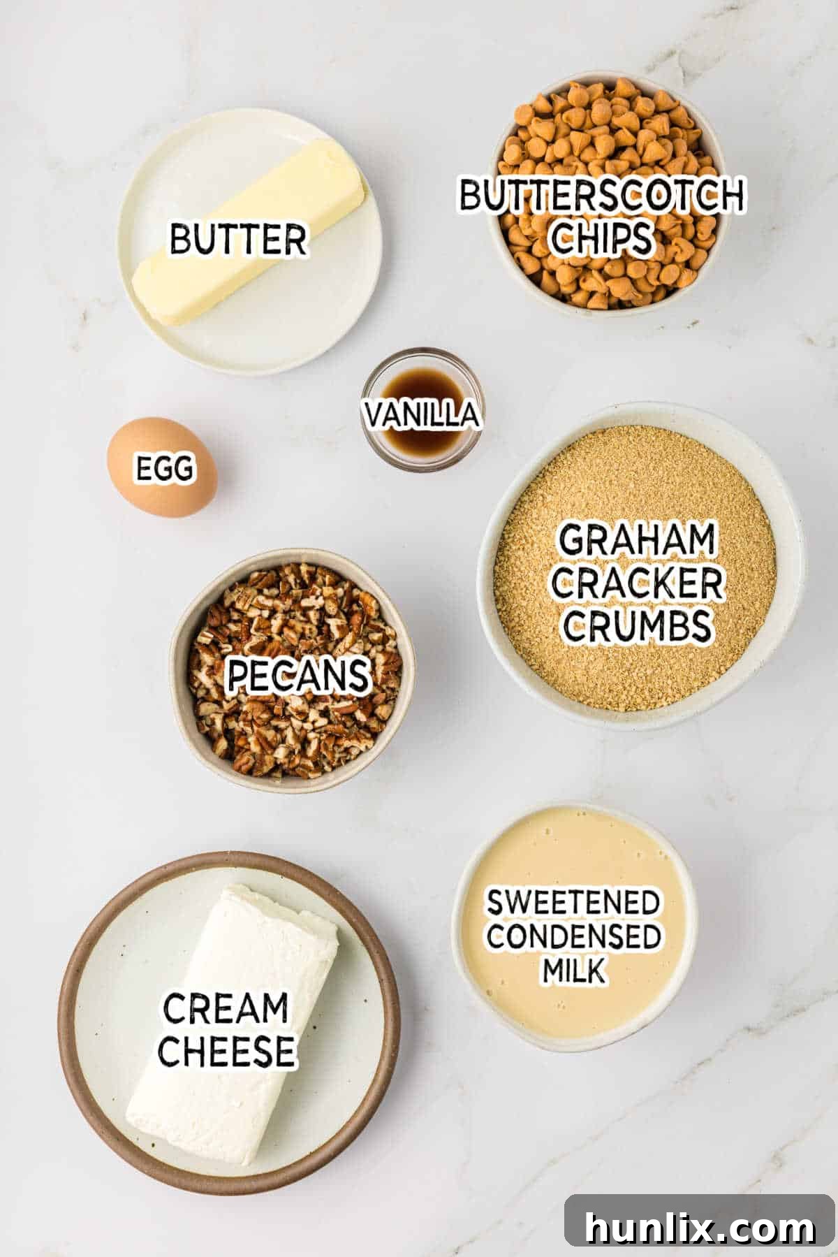 Dreamy Butterscotch Cheesecake Squares 3 A selection of ingredients for butterscotch cheesecake bars, including butterscotch chips, butter, graham cracker crumbs, pecans, cream cheese, sweetened condensed milk, vanilla extract, and an egg, arranged on a white surface.