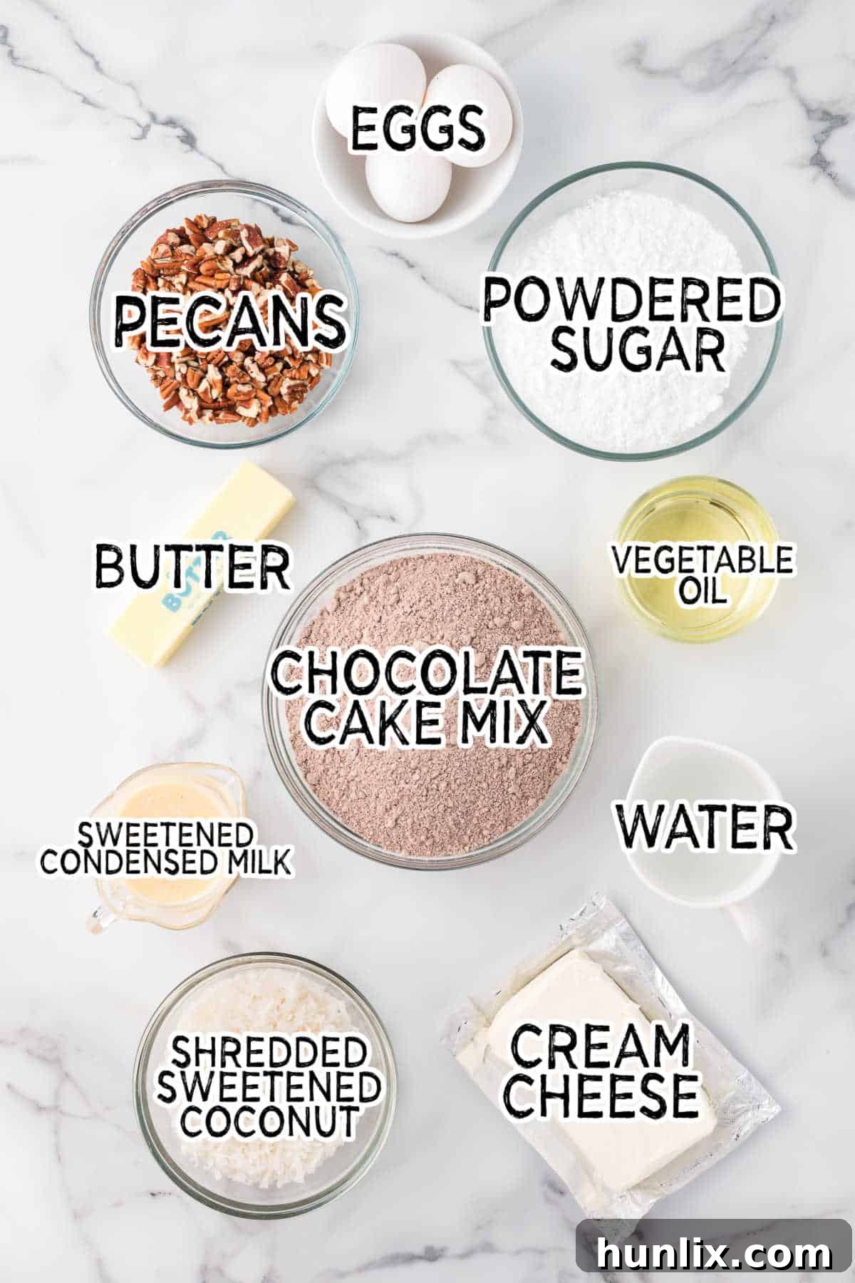 Various baking ingredients including cream cheese, butter, sugar, pecans, coconut, chocolate cake mix, and eggs on a white marble background.