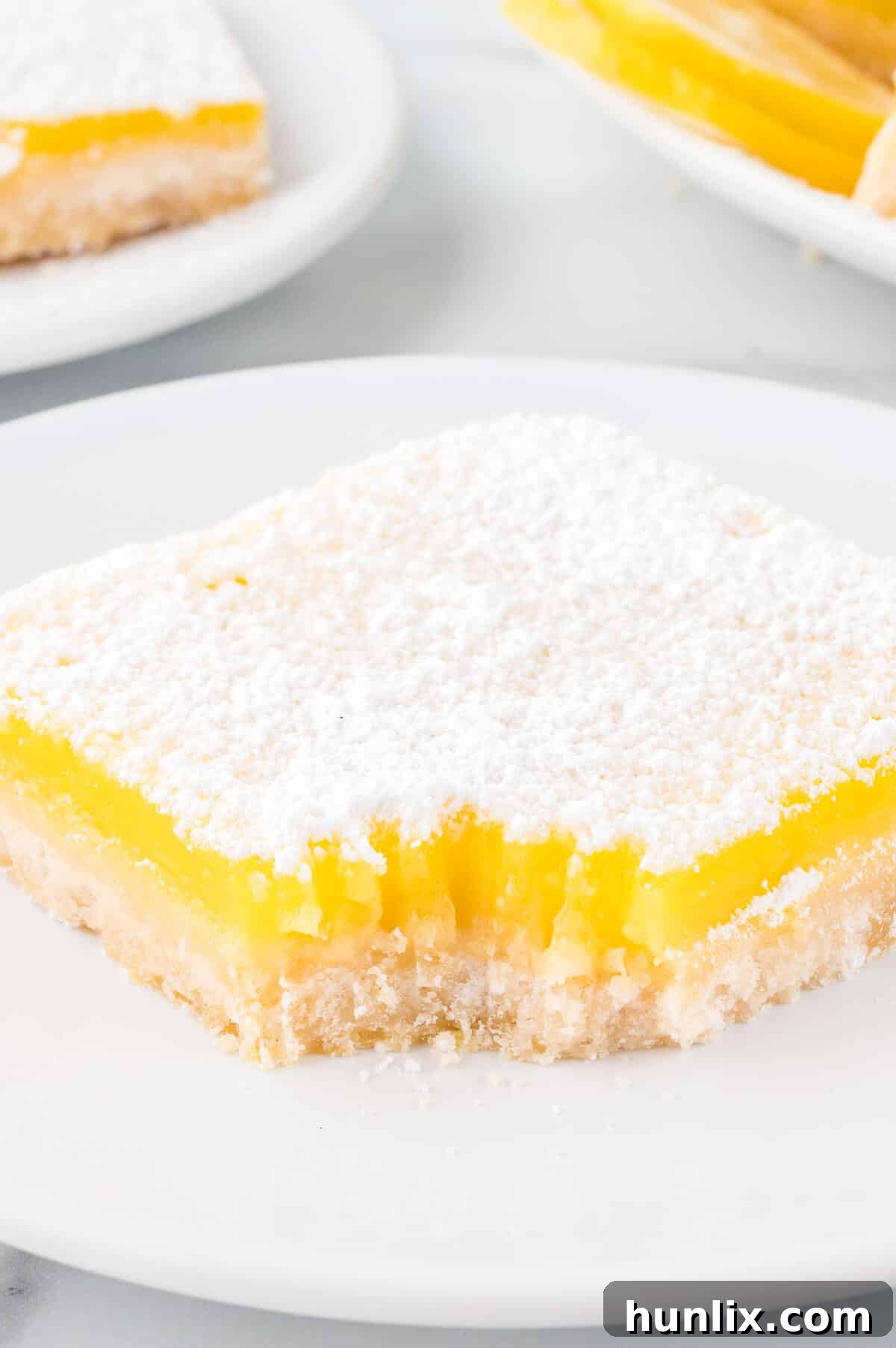 A lemon bar on a plate with a bite taken out of it, showcasing the filling and crust.