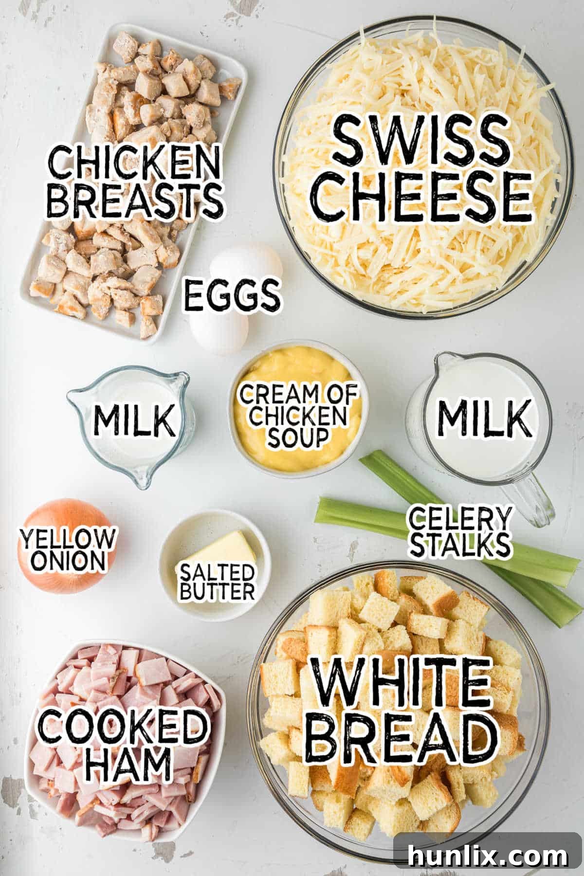 A collection of fresh ingredients laid out on a kitchen counter, including celery, onion, butter, eggs, milk, white bread cubes, diced cooked ham, cooked chicken, shredded Swiss cheese, and a can of cream of chicken soup.
