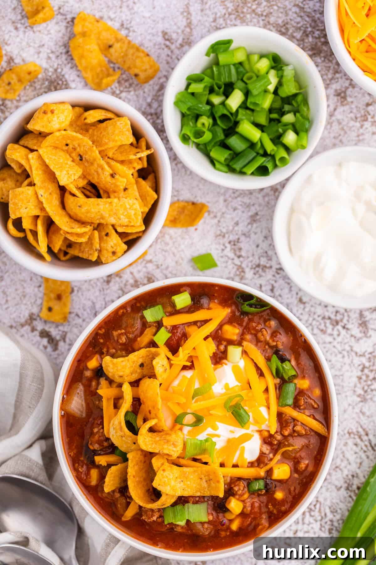 A white bowl of taco chili topped with shredded cheese, sour cream, and green onions, with corn chips on the side.