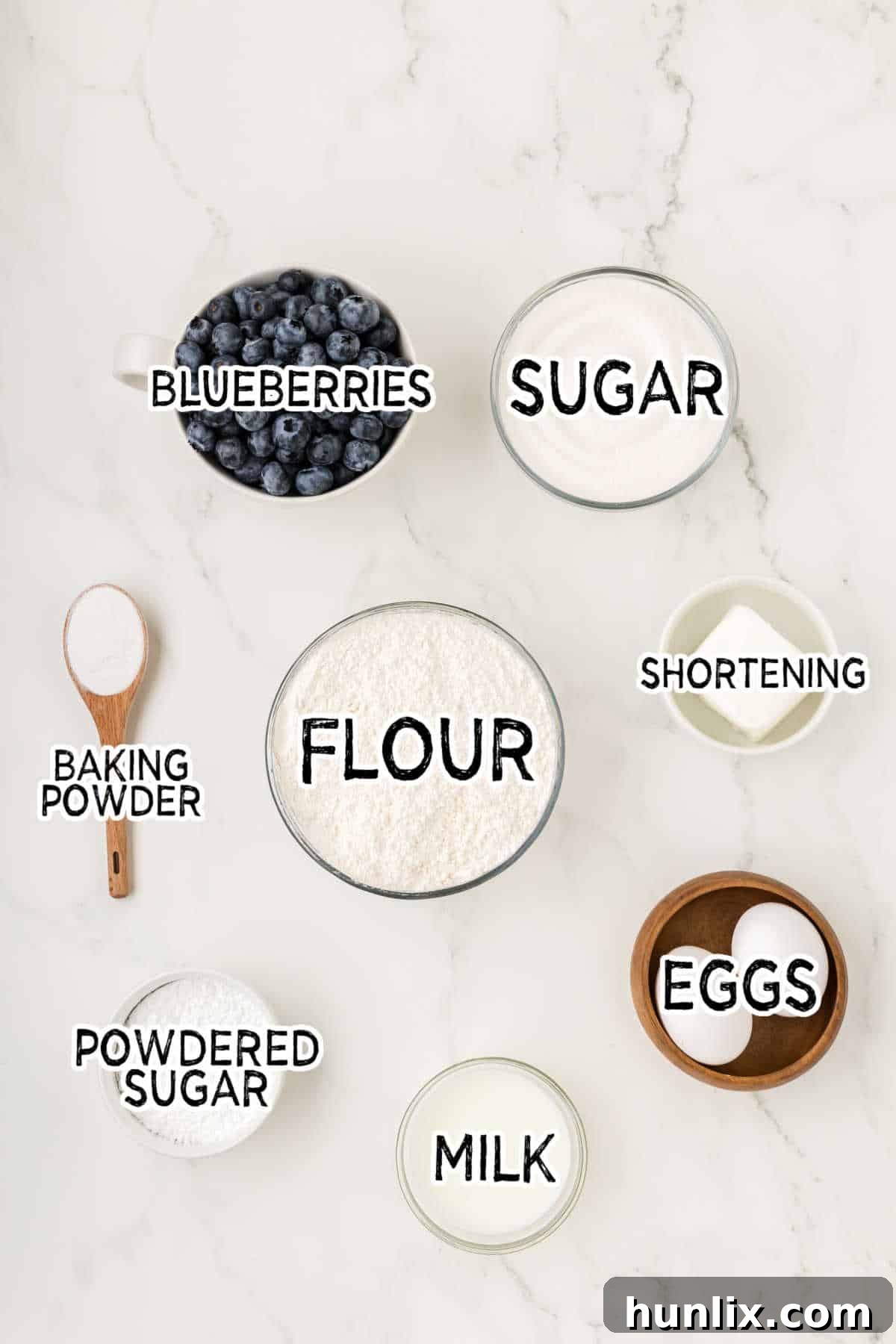 A selection of fresh ingredients including shortening, sugar, eggs, flour, milk, and blueberries laid out on a white marble background.