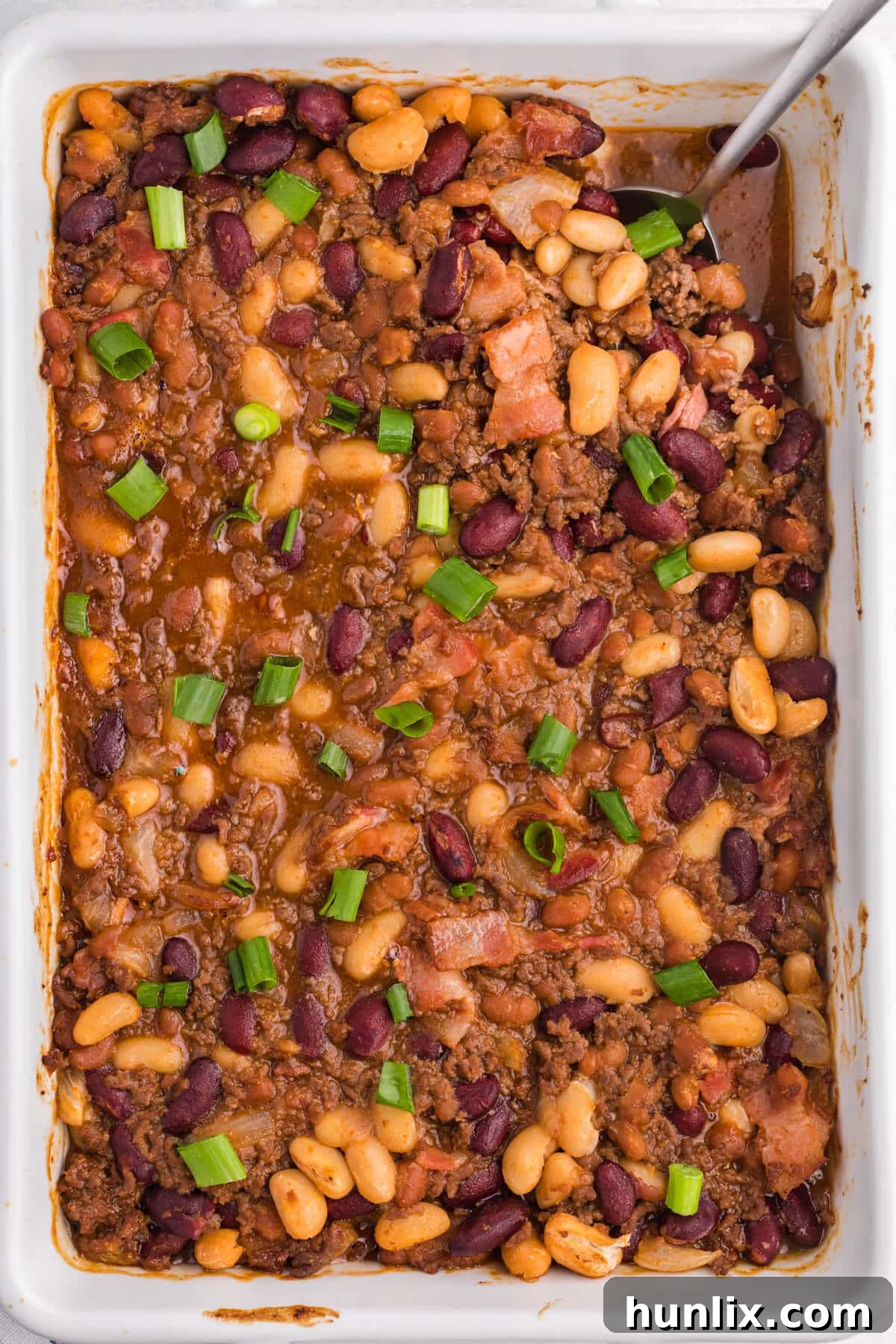 A baked bean casserole served in a white dish with a large serving spoon.