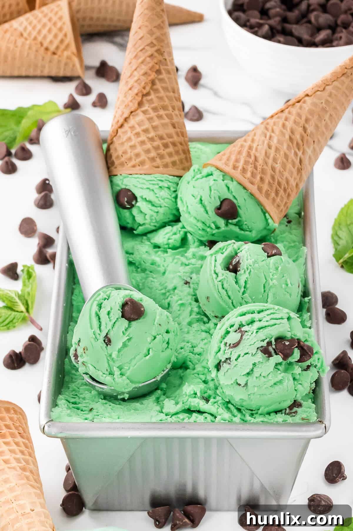 A loaf pan filled with mint chocolate chip ice cream, alongside an ice cream scoop and two waffle cones.