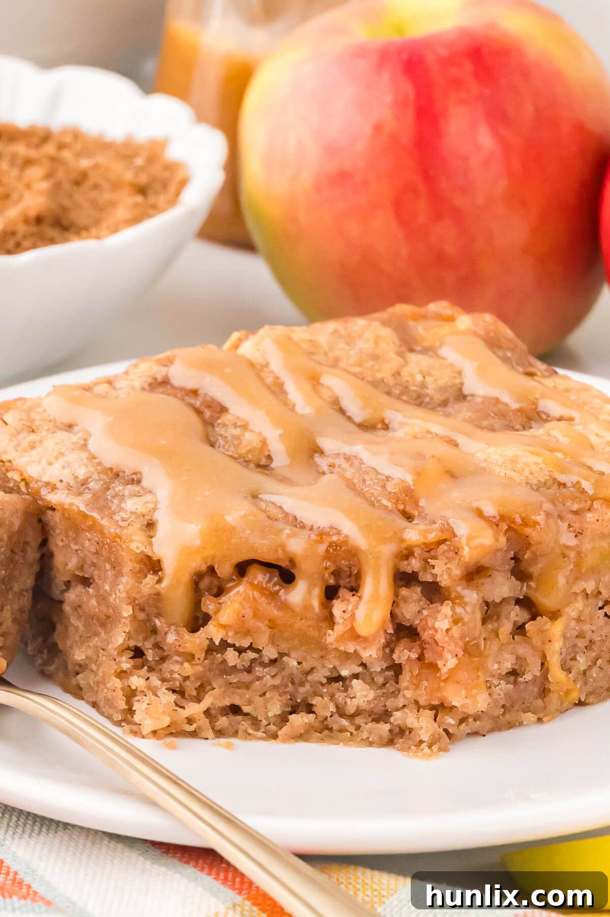 A close-up shot of a single slice of apple cake on a plate, with a bite taken from the end, revealing the moist interior and chunks of apple, all covered in butterscotch sauce.