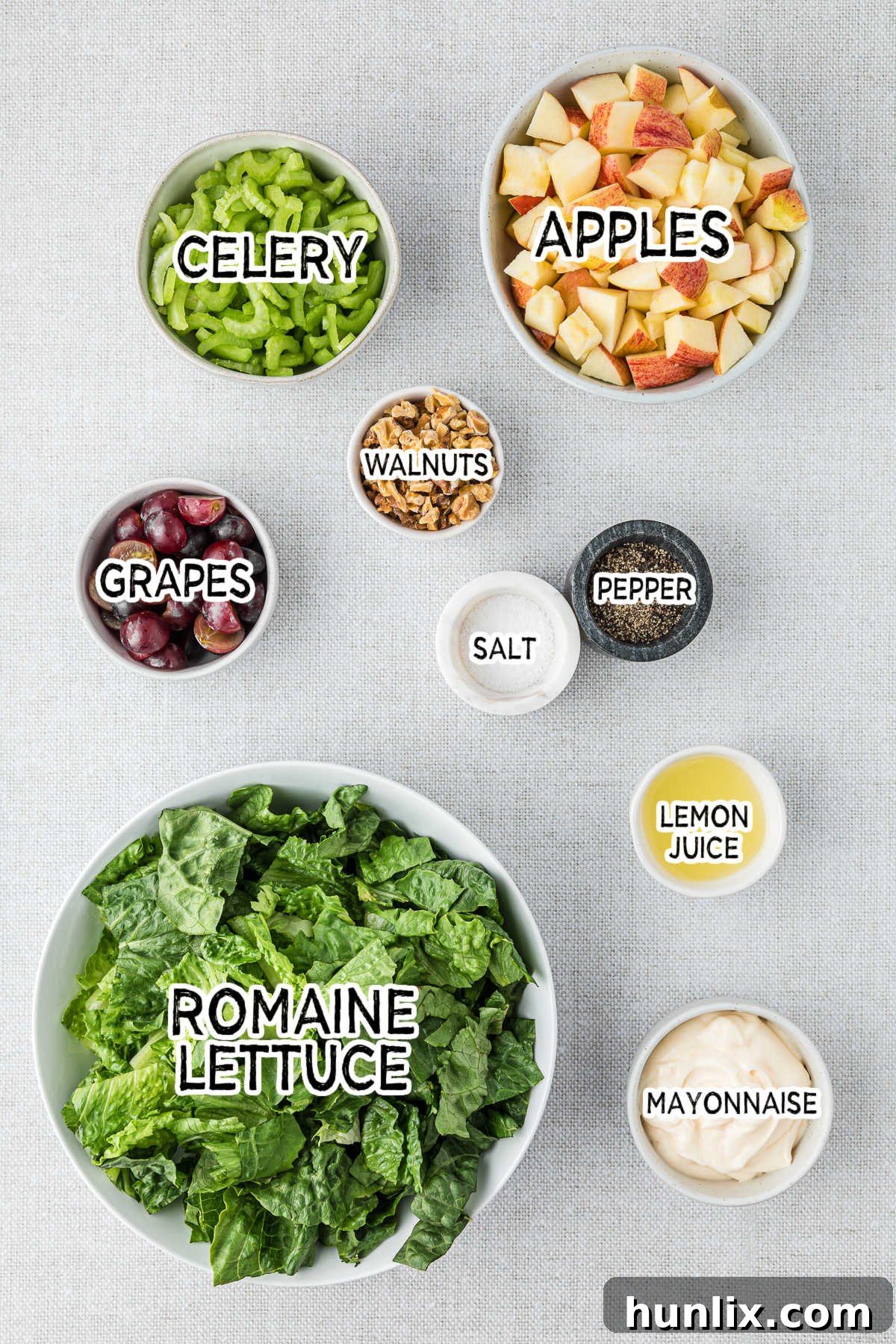A close-up view of the fresh ingredients for Waldorf salad laid out on a wooden surface, including a bowl of chopped apples, sliced celery, red grapes, and walnuts, ready for preparation.
